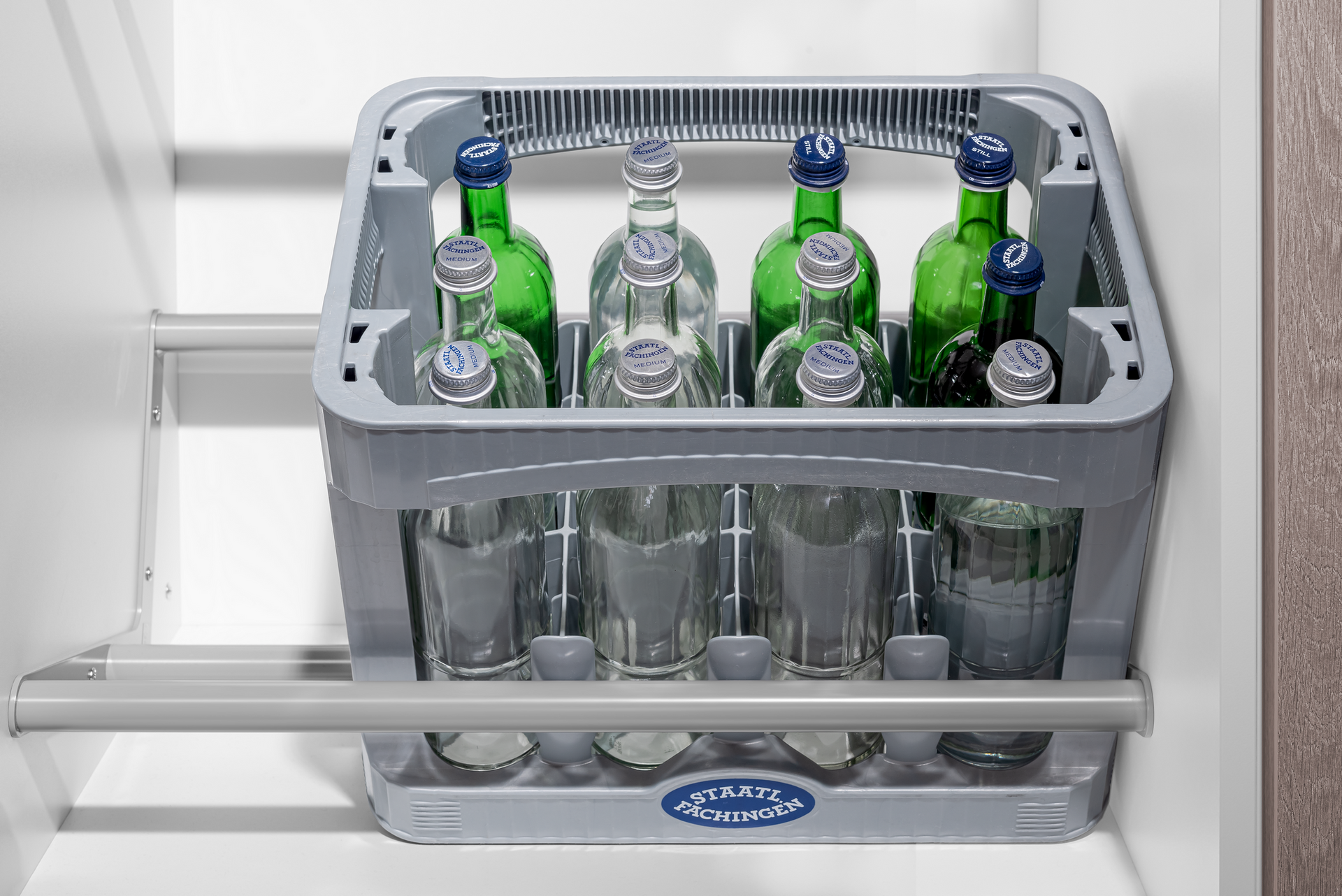A grey bottle crate filled with glass bottles, pulled out from a cabinet drawer. Some bottles are green.