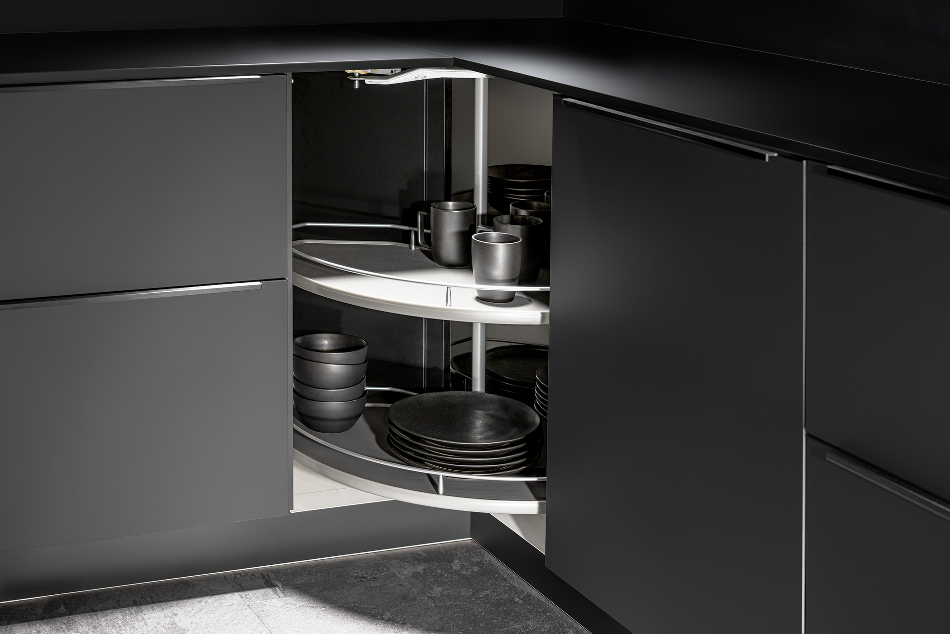 A dark gray corner kitchen cabinet with a rotating shelf containing dark dishes and mugs.