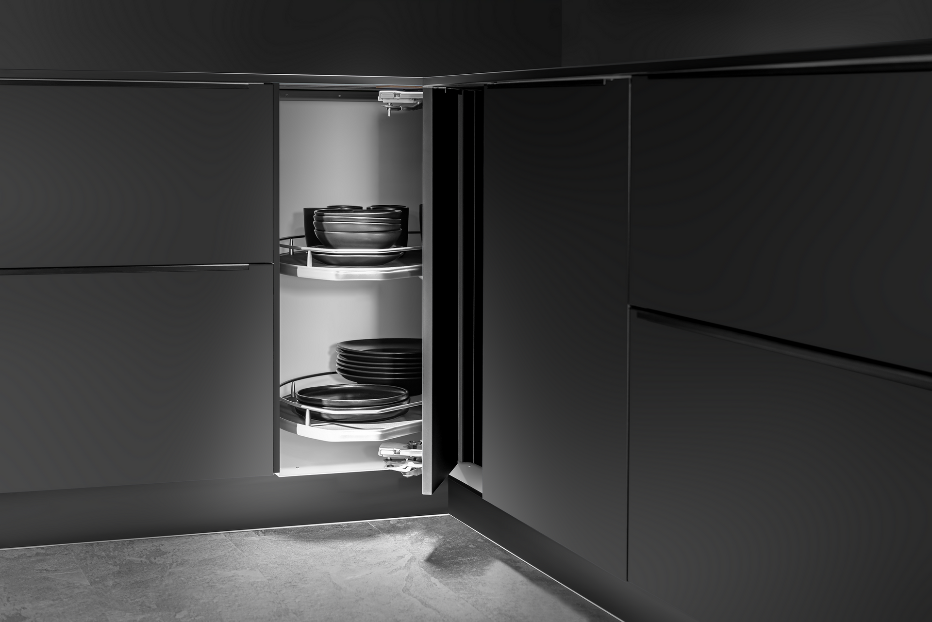 A kitchen cabinet corner with a pull-out, two-tiered carousel for storage, filled with pots and pans. Black cabinetry, grayscale.