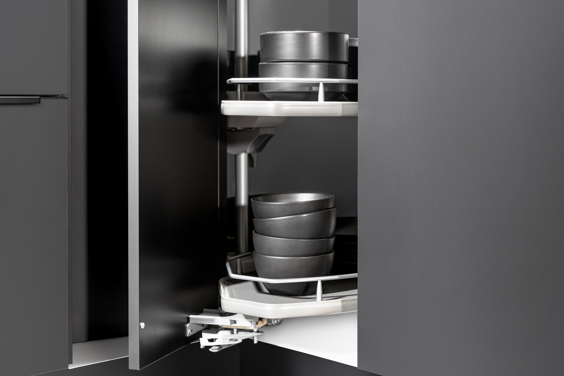 A gray kitchen cabinet with pull-out, rotating shelves holding stacks of dark bowls and pans.