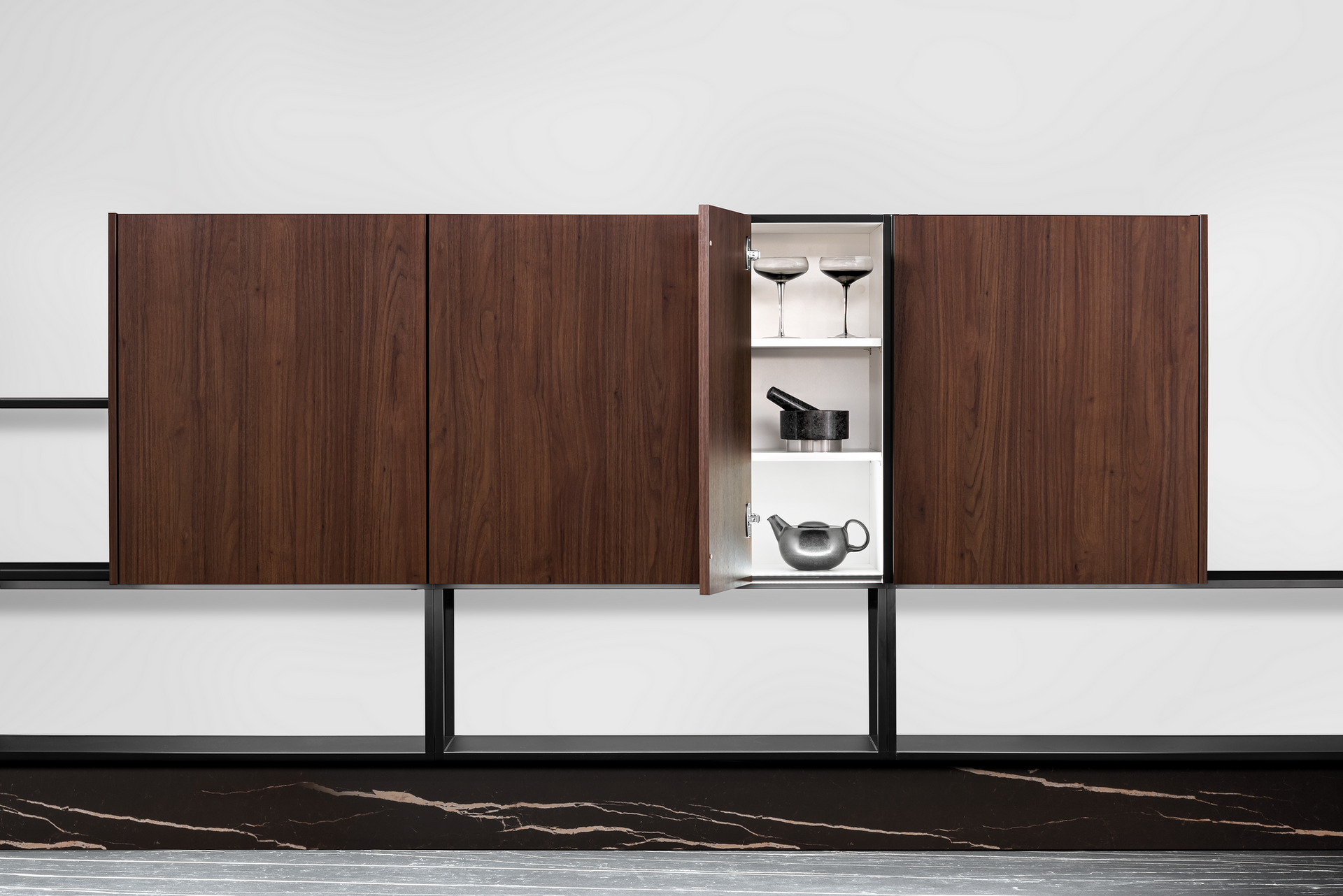 Wooden wall-mounted cabinet with a door open, revealing glassware and utensils. Black metal supports frame the cabinet against a white wall.