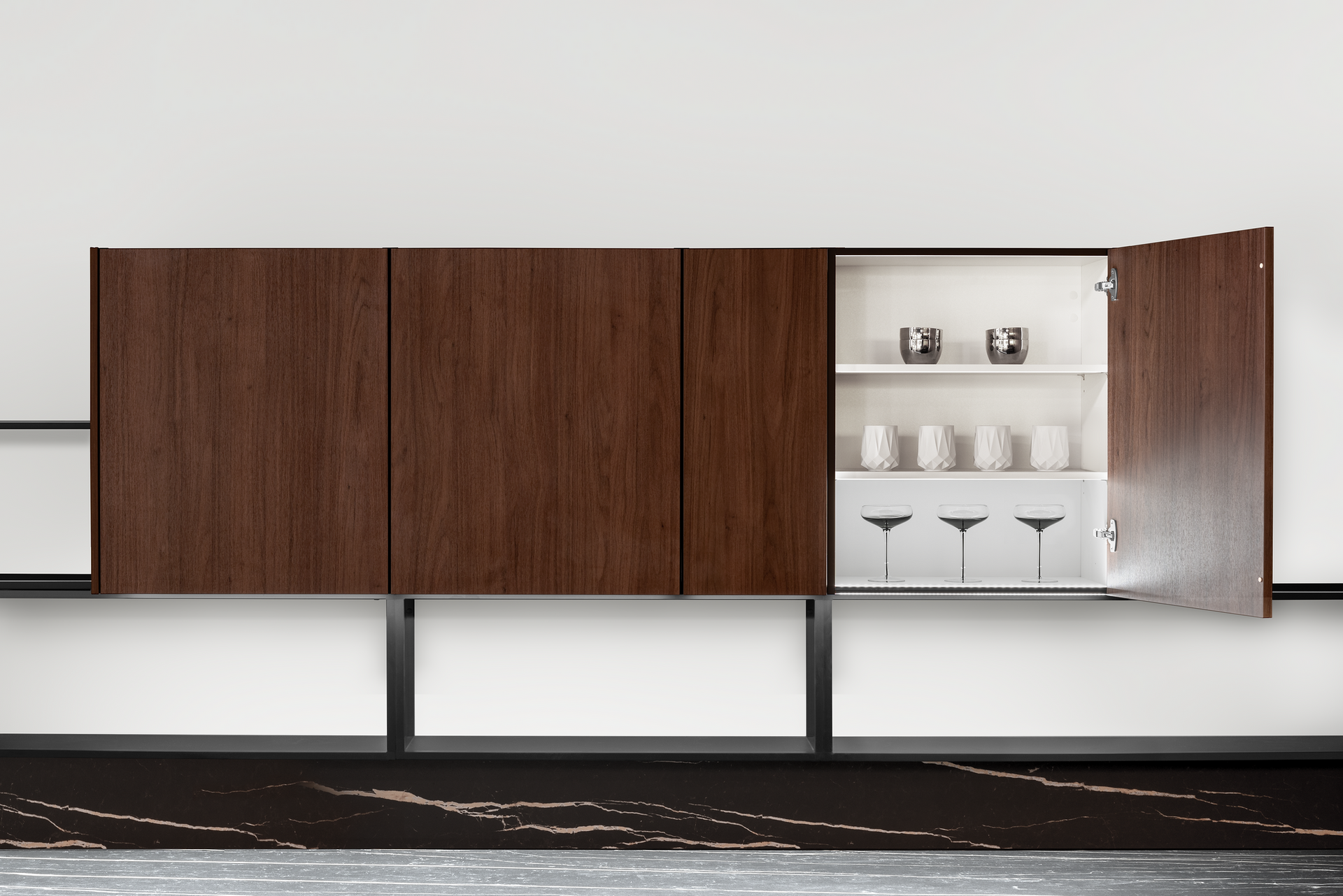 A wall-mounted cabinet with three dark wood doors, one open to reveal white interior shelves holding glasses and cups.