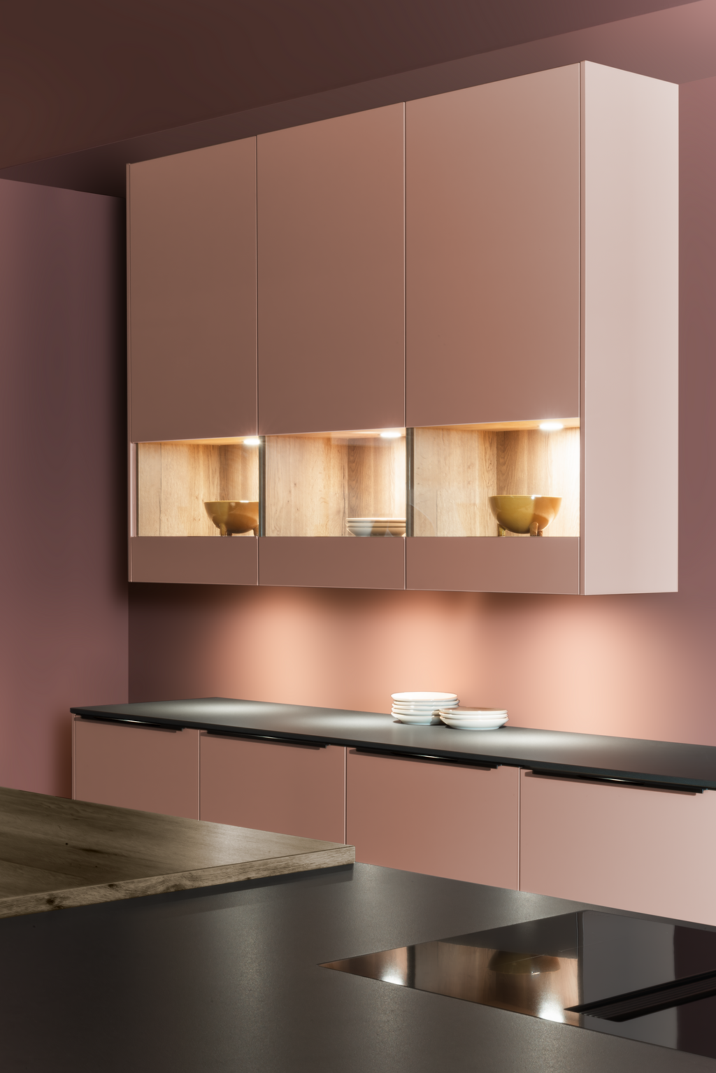 Modern kitchen with pink cabinets, light wood shelving, and dark countertops. Warm lighting illuminates the space.