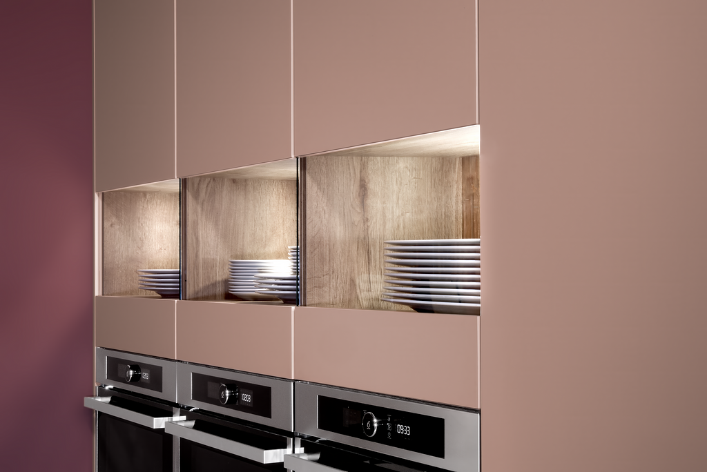 Built-in kitchen cabinetry with recessed, illuminated shelves displaying dishes and three oven appliances.