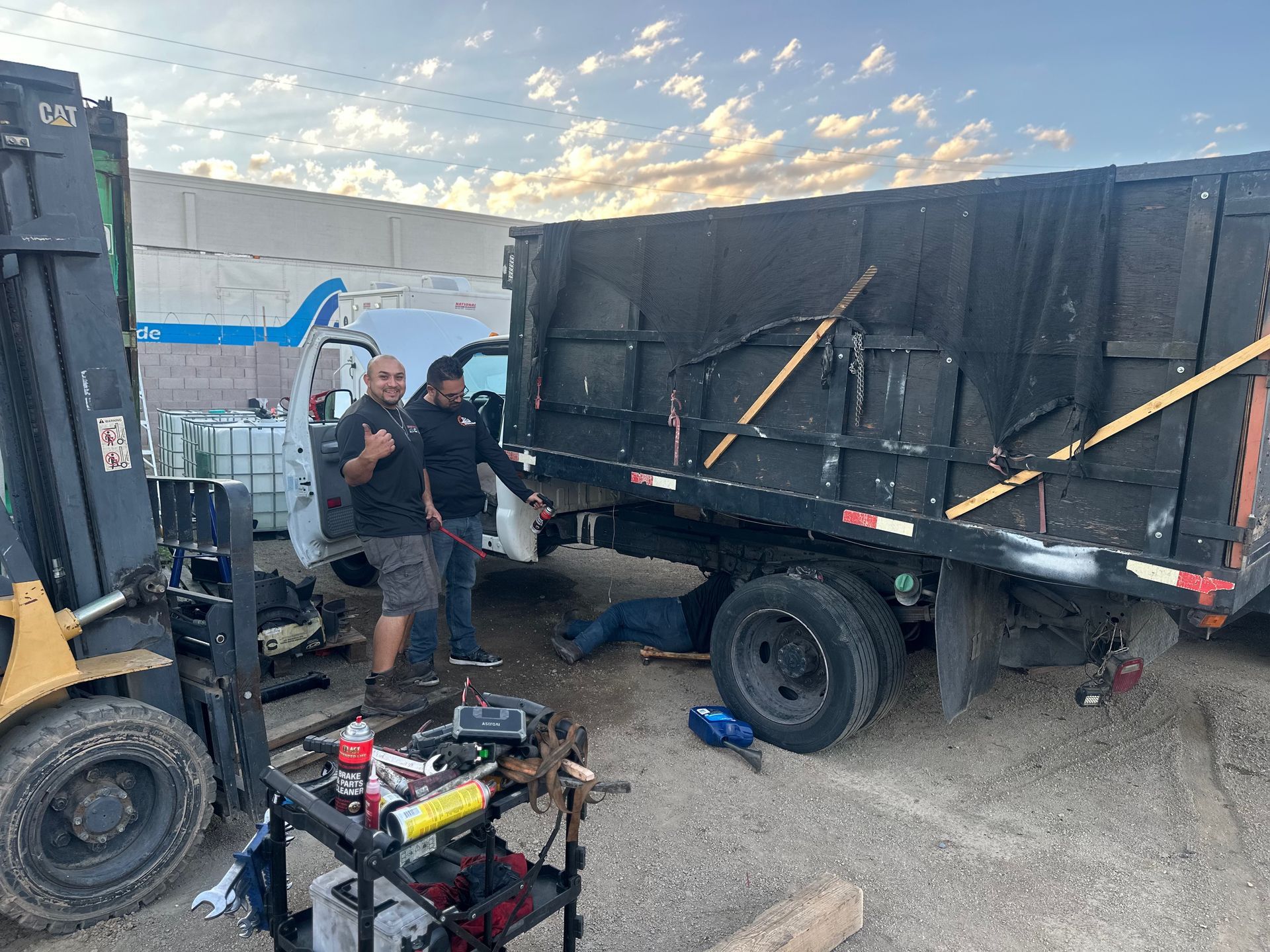 Two men stand by a truck while another works underneath. Tools on a cart, forklift present.