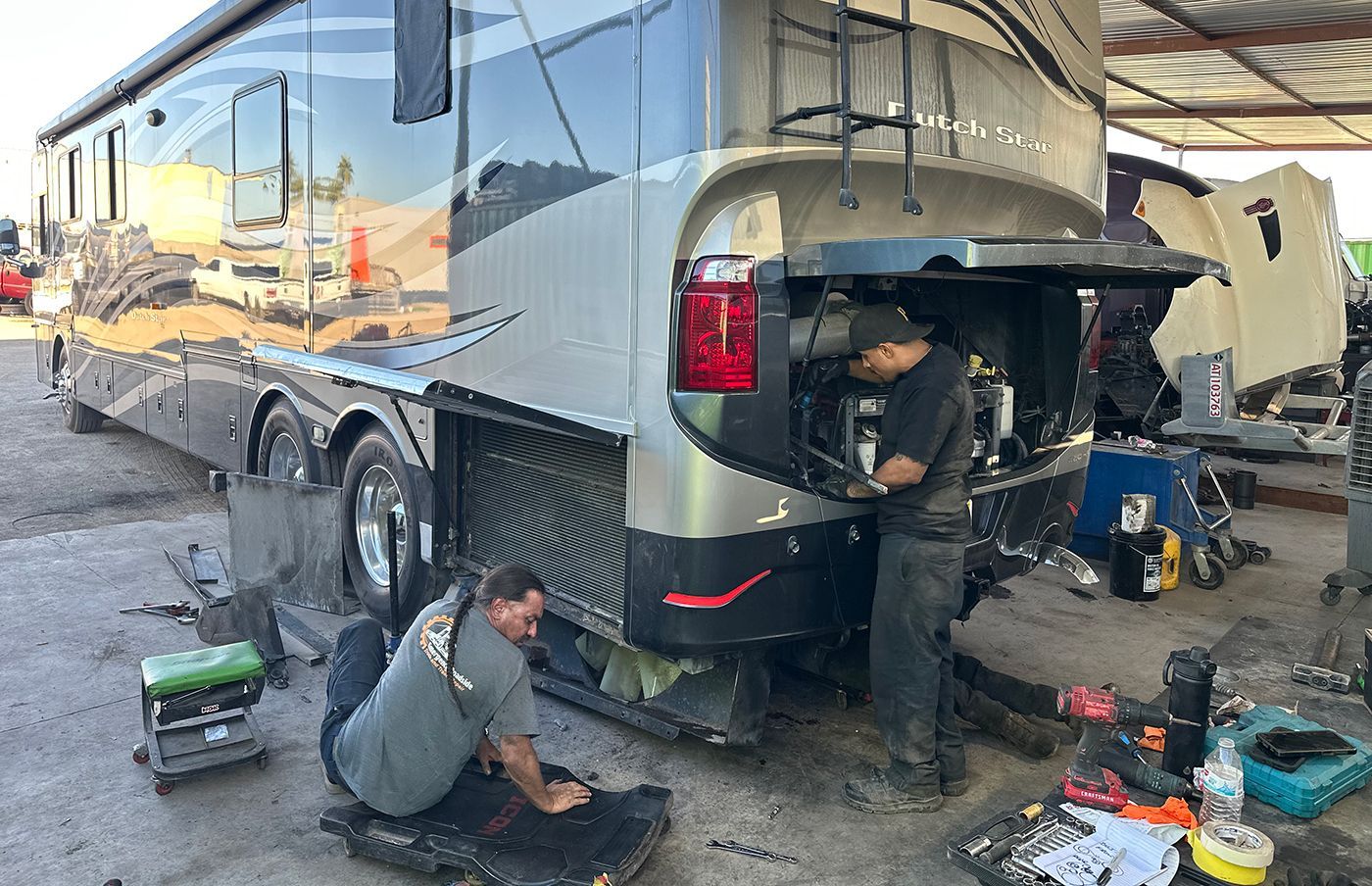 Two mechanics working on the back of an RV in a repair shop; tools and equipment scattered.