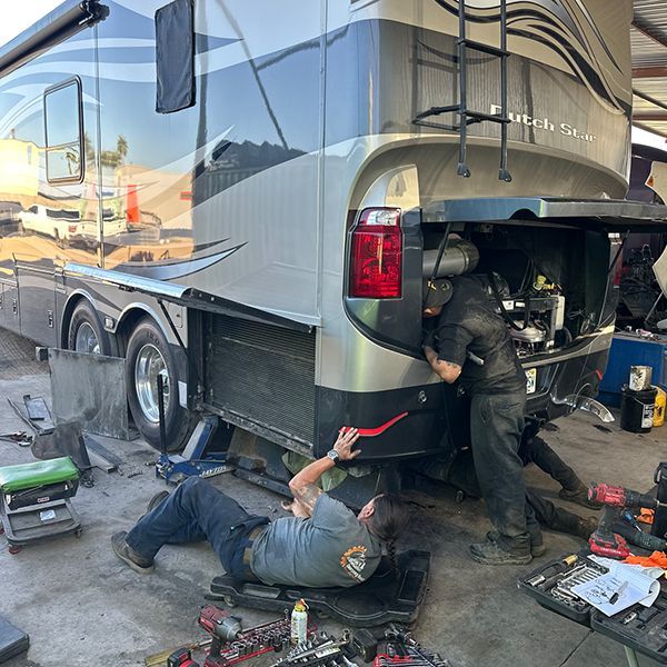 Two mechanics work on the engine of a Dutch Star RV at a repair shop; one is underneath the vehicle.