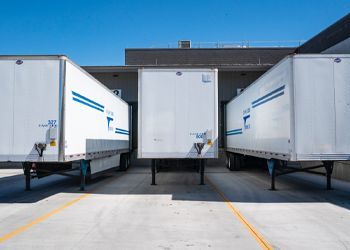 Three white semi trucks are parked in a parking lot in front of a building.