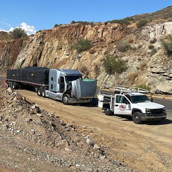 Silver semi-truck with open hood next to a white service truck on a rocky road in front of a hillside.
