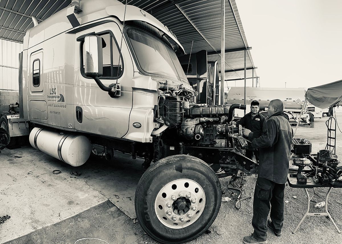 Truck with open hood, being worked on by two people outdoors under a shelter.
