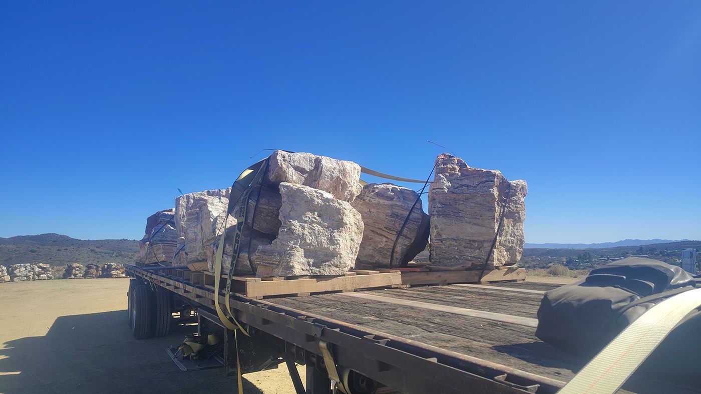 Flatbed truck carrying large, white-wrapped rocks secured with straps under a clear blue sky.