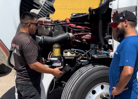 Two men examining a truck engine. One holds a part. Open hood. Outdoors, sunny.
