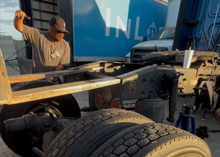 Man working on the undercarriage of a semi-truck; outdoors, day. Truck has blue cab, INL logo.