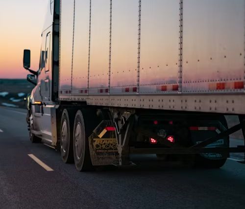 Semi-truck driving on a road at dusk, with a pink and orange sky.
