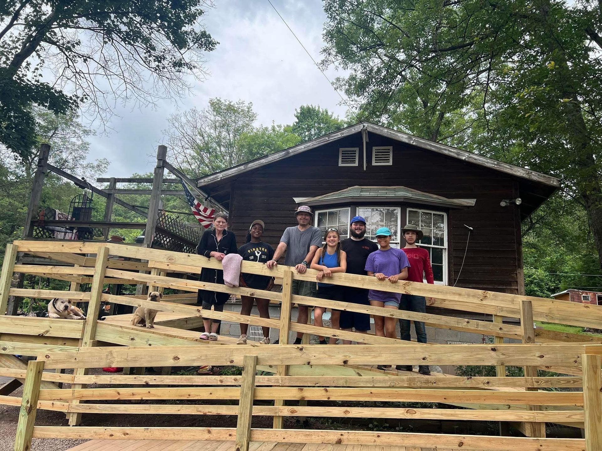 Group of people smiling on a wooden ramp in front of a house. Overcast day, trees in background.