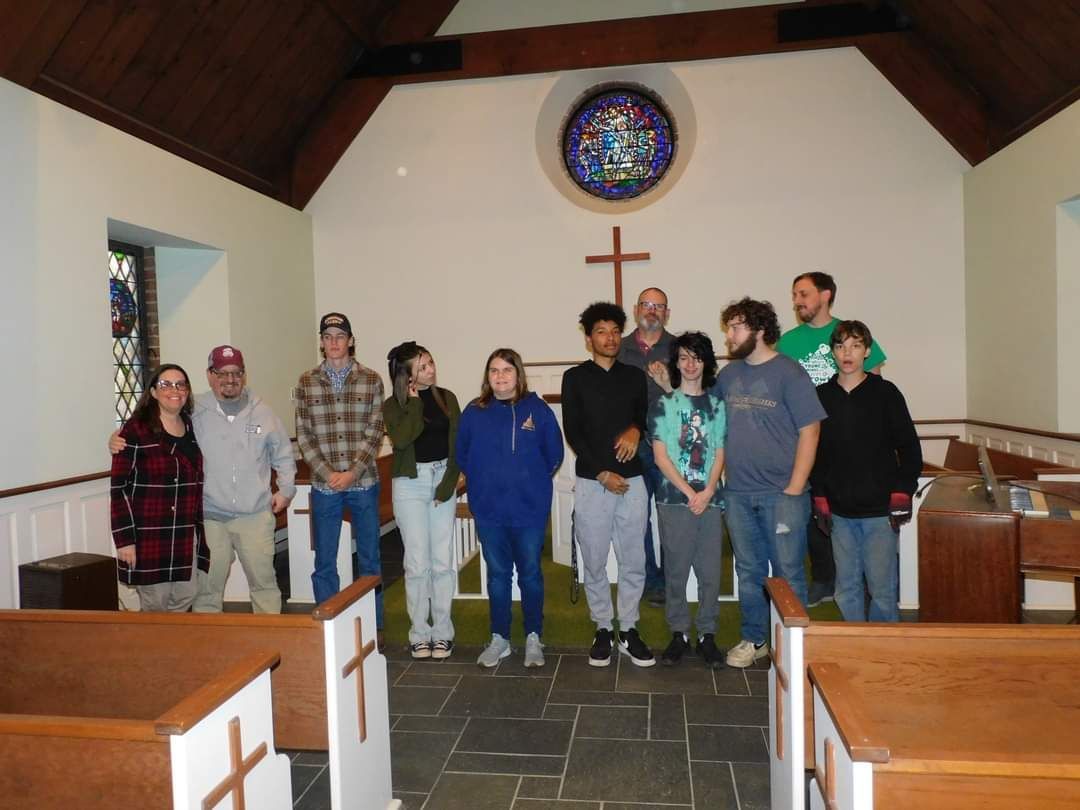 Group of people inside a church, standing in front of a cross and stained-glass window.