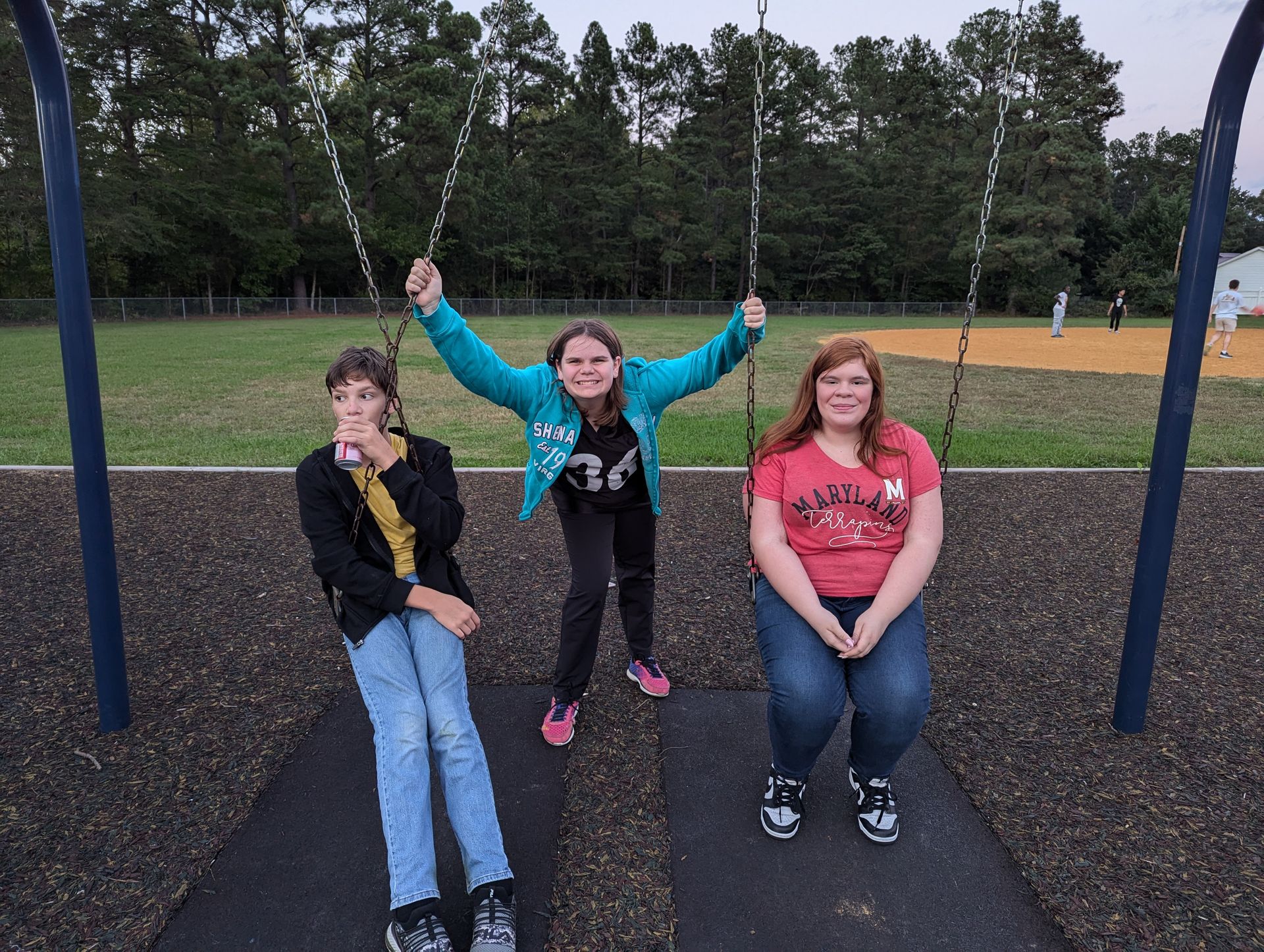Three girls on swings at a park; one girl raises arms with a smile. Dusk setting.