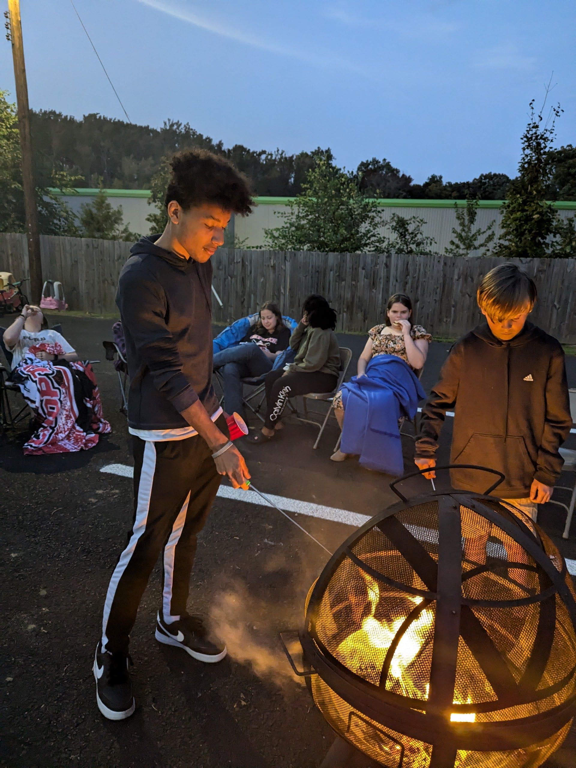 Group of people around a fire pit at dusk. A young man stirs the flames. Others sit nearby in chairs.