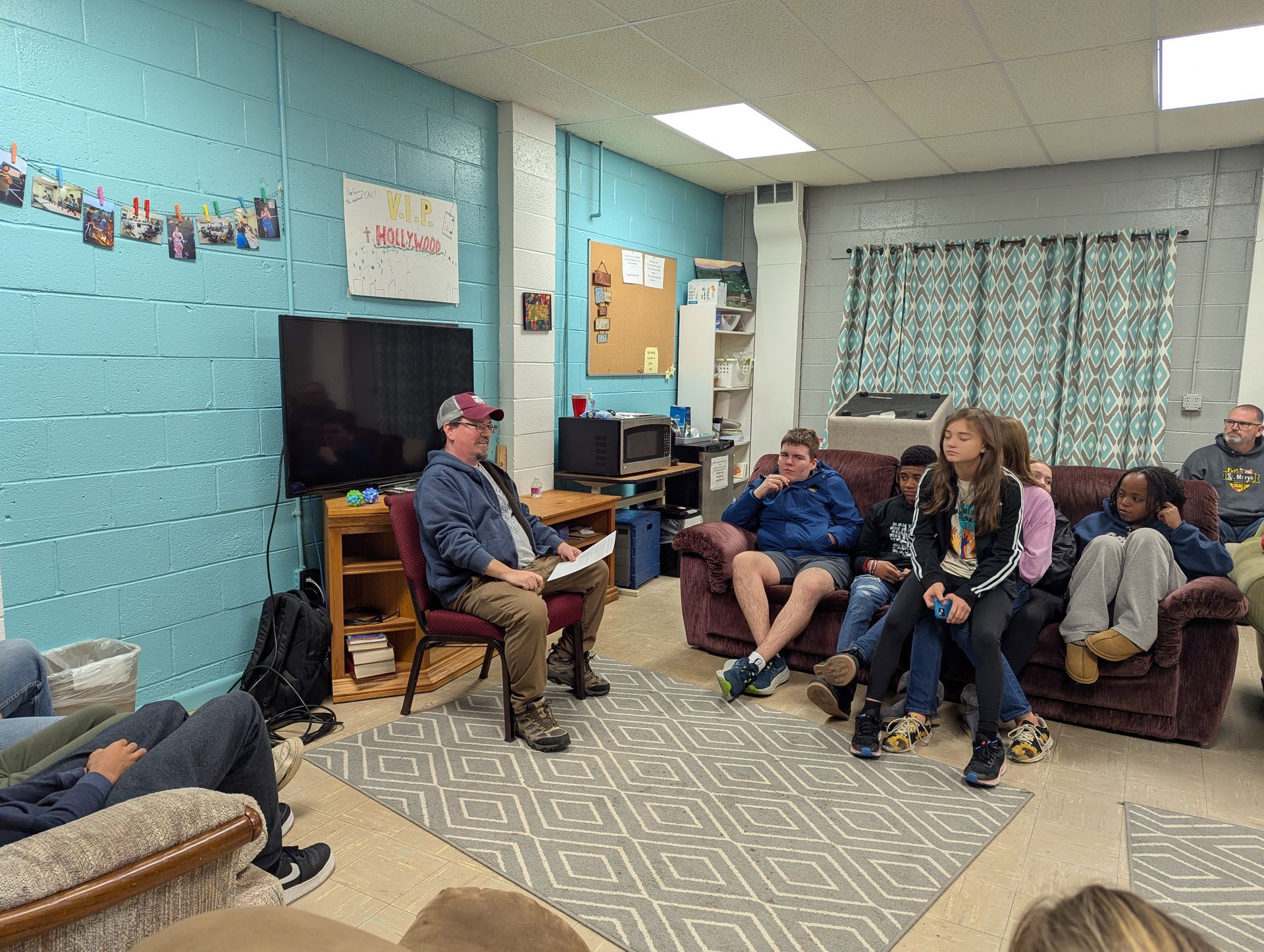 A group of people in a room listening to a man speaking. Blue walls, couch, and television are visible.