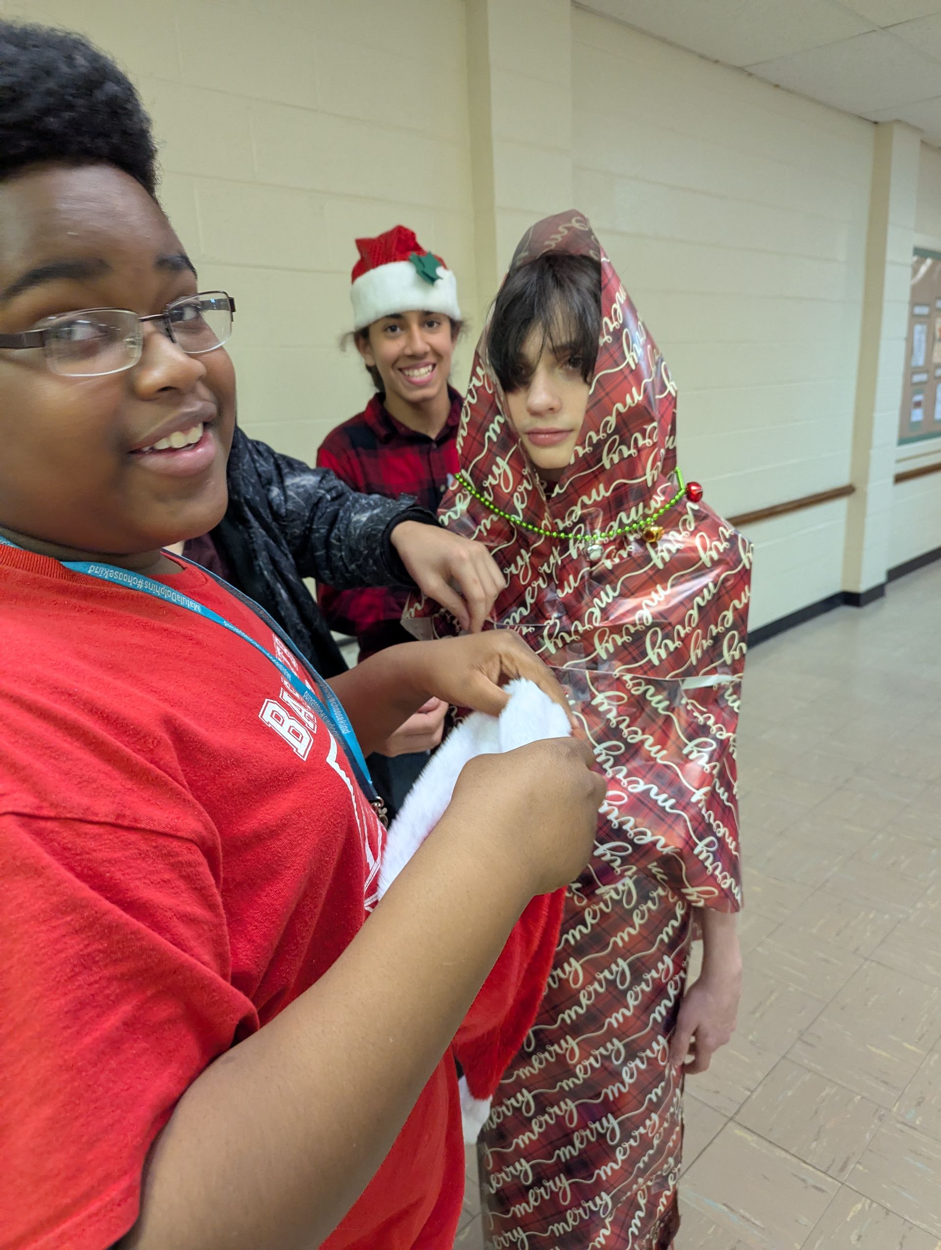 Three students: one in a red shirt is wrapping another, who is in gift wrap, while another wears a Santa hat.