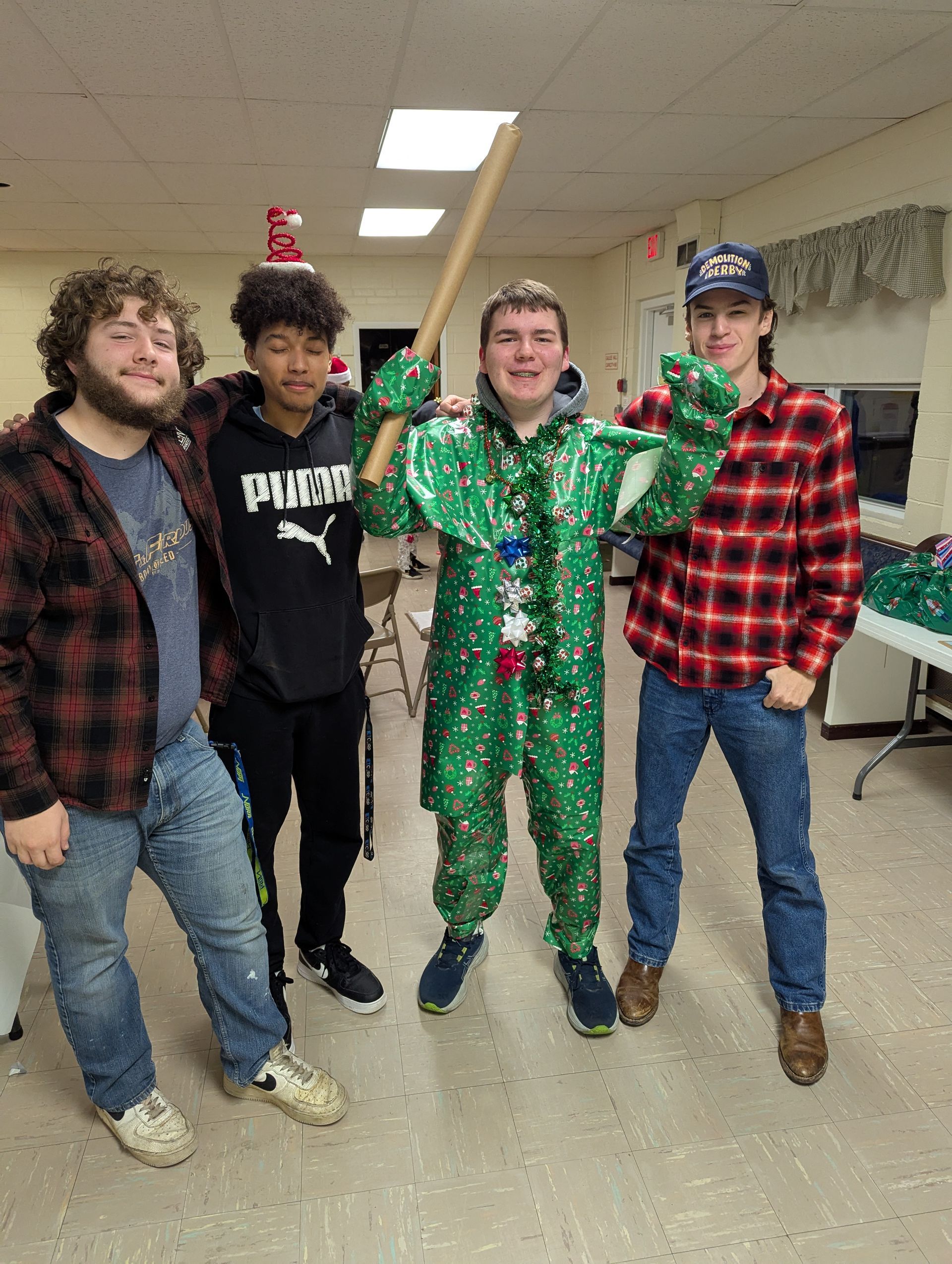 Four young men pose indoors, one in a green Christmas onesie holding a wooden bat, others in casual attire.