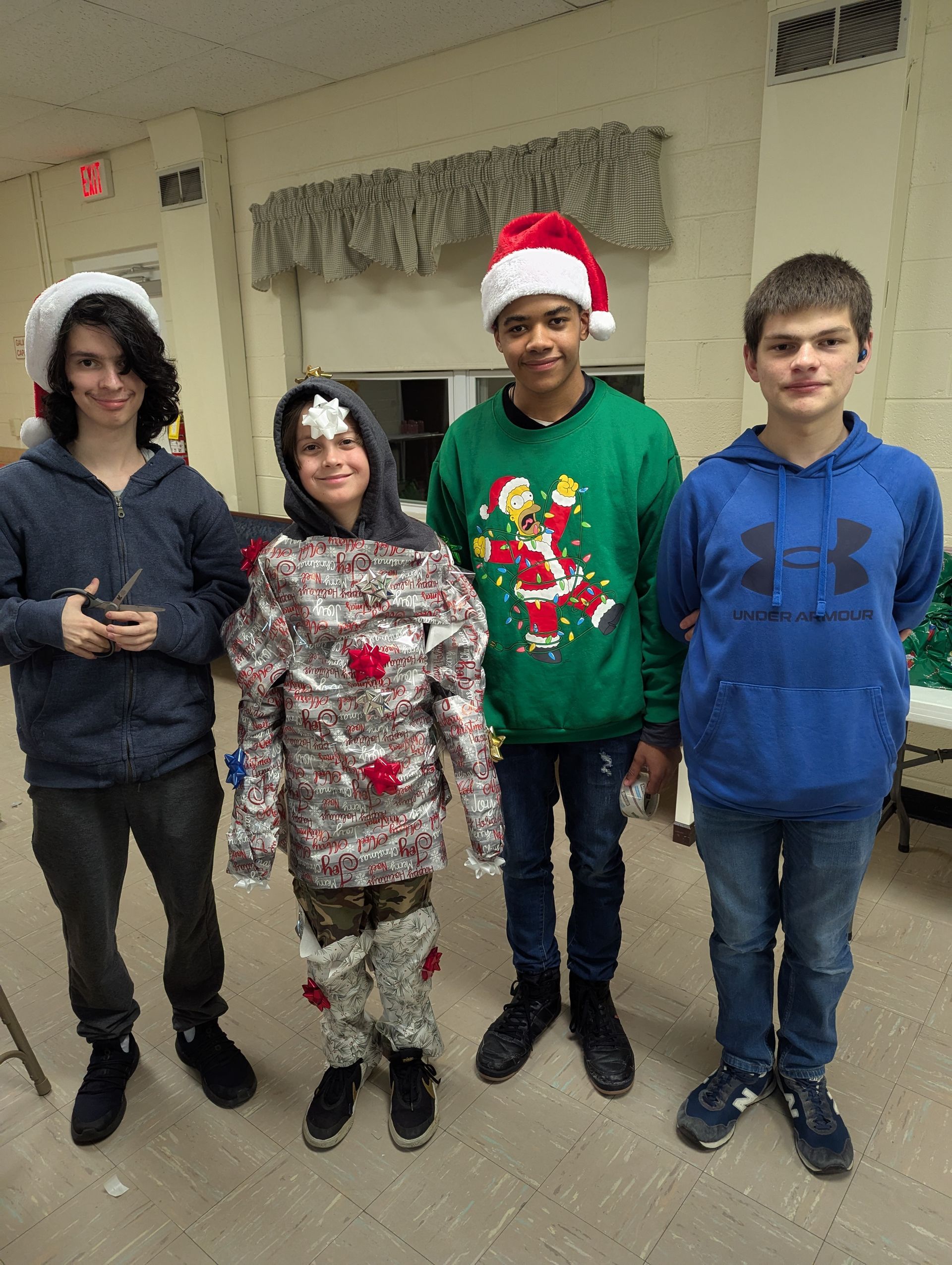 Four young people in festive attire pose indoors. One wears a Santa hat and Christmas sweater.