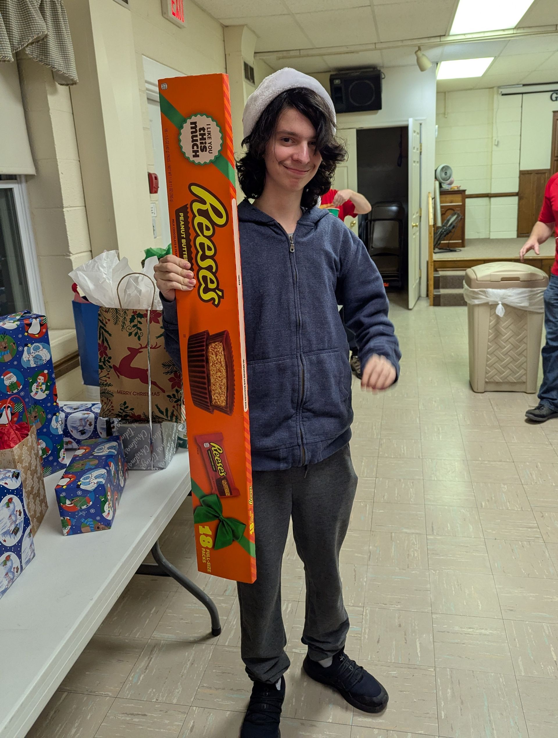 Person holding a giant Reese's candy bar, wearing a Santa hat, smiling. Surrounded by gifts.