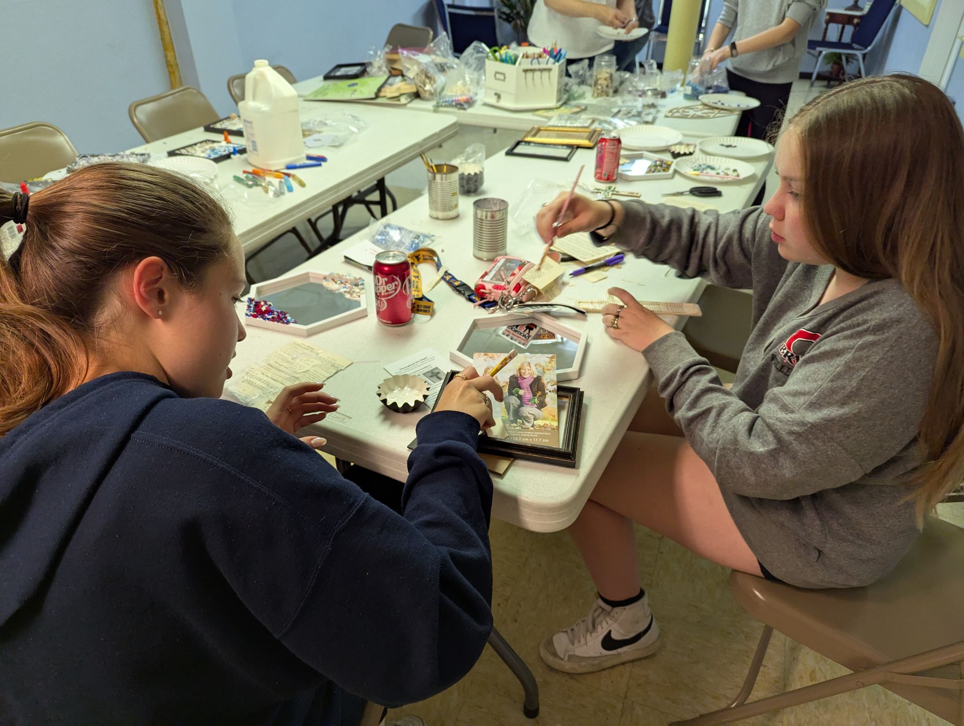 Two girls crafting at a table, painting and working on small objects. Indoor setting, bright.