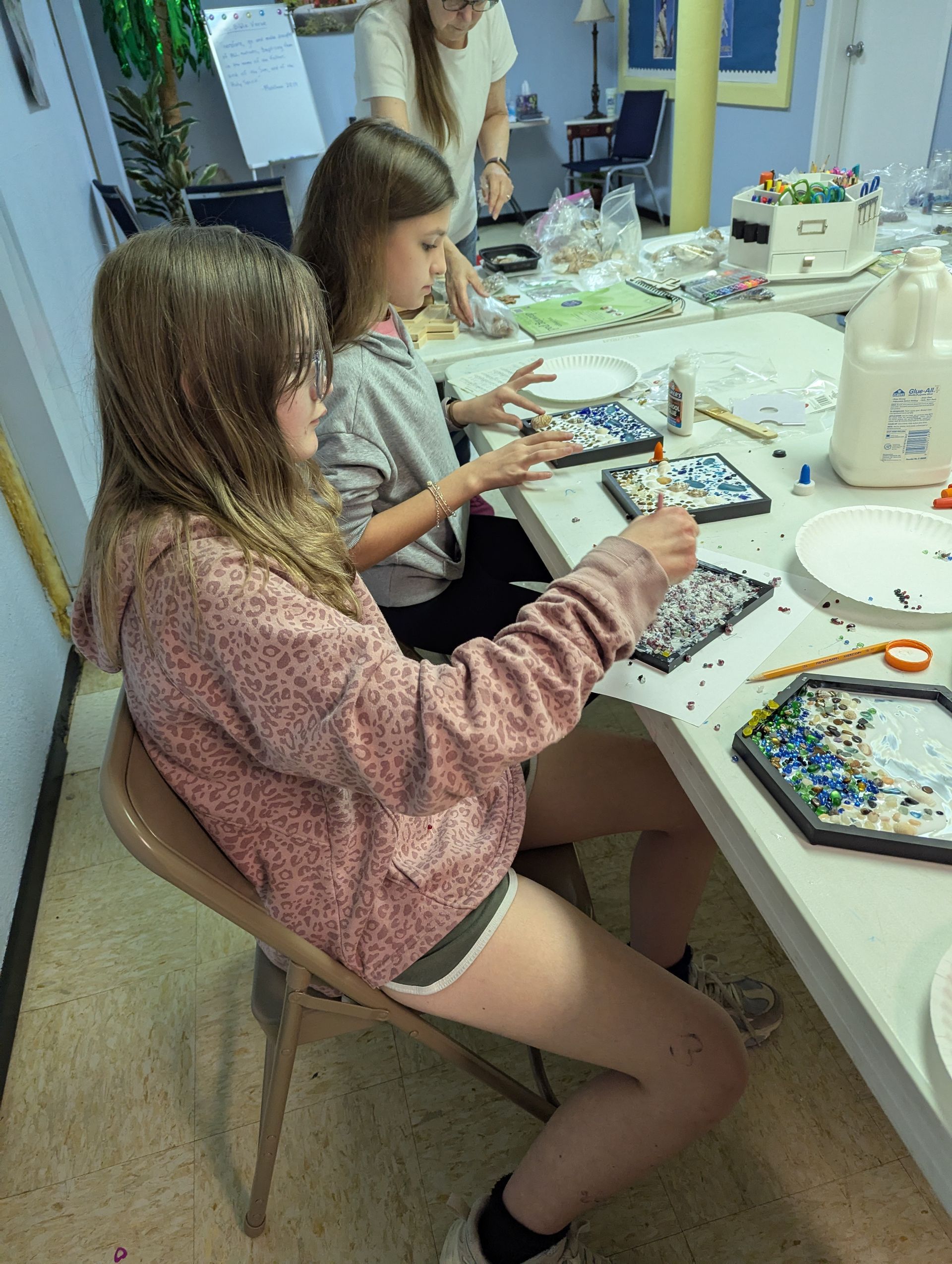 Two girls crafting at a table, applying small pieces to black tiles. Another person stands behind them.