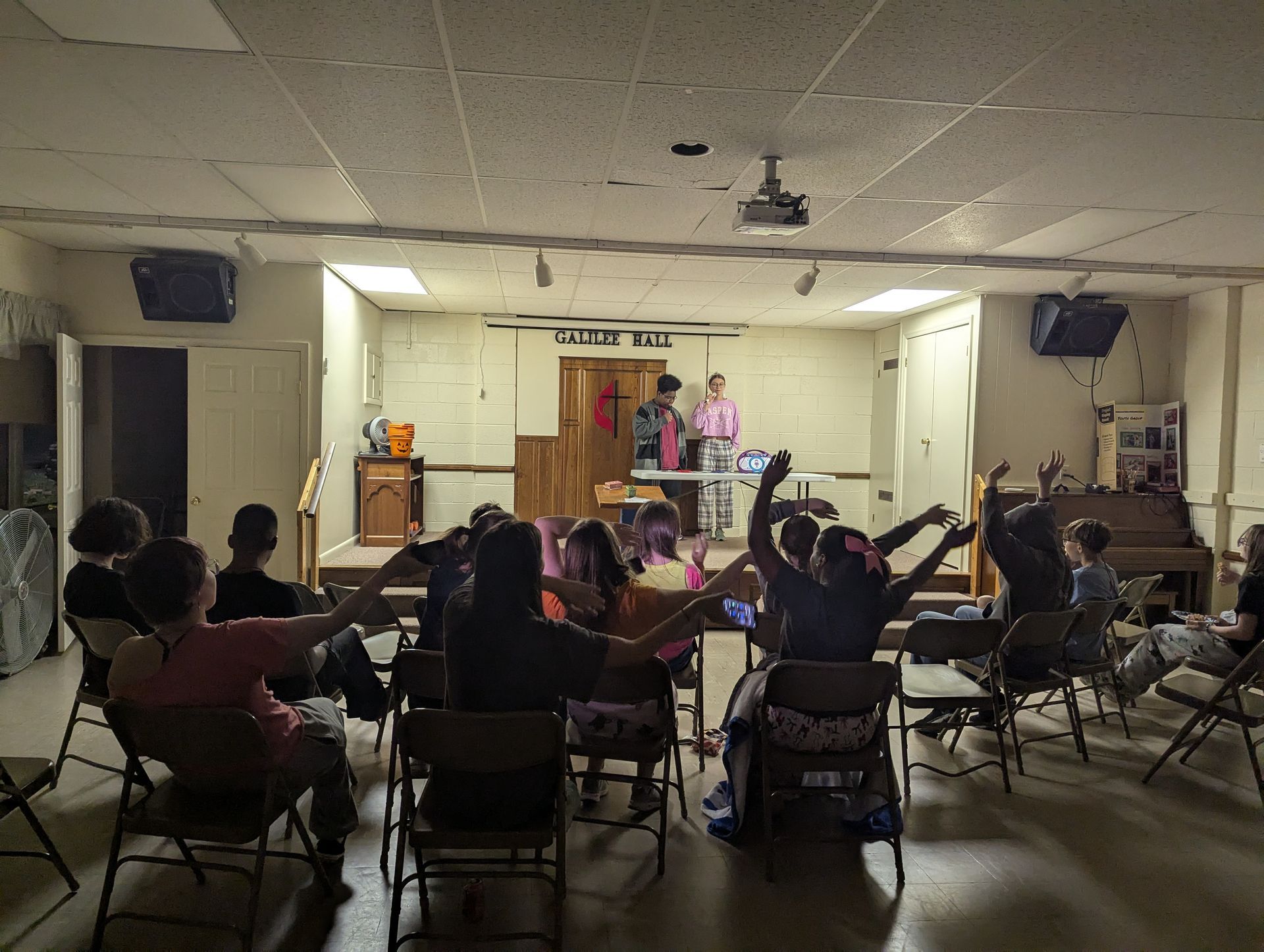 People in a room with arms raised, facing a stage. Two people are on the stage; one plays a drum.