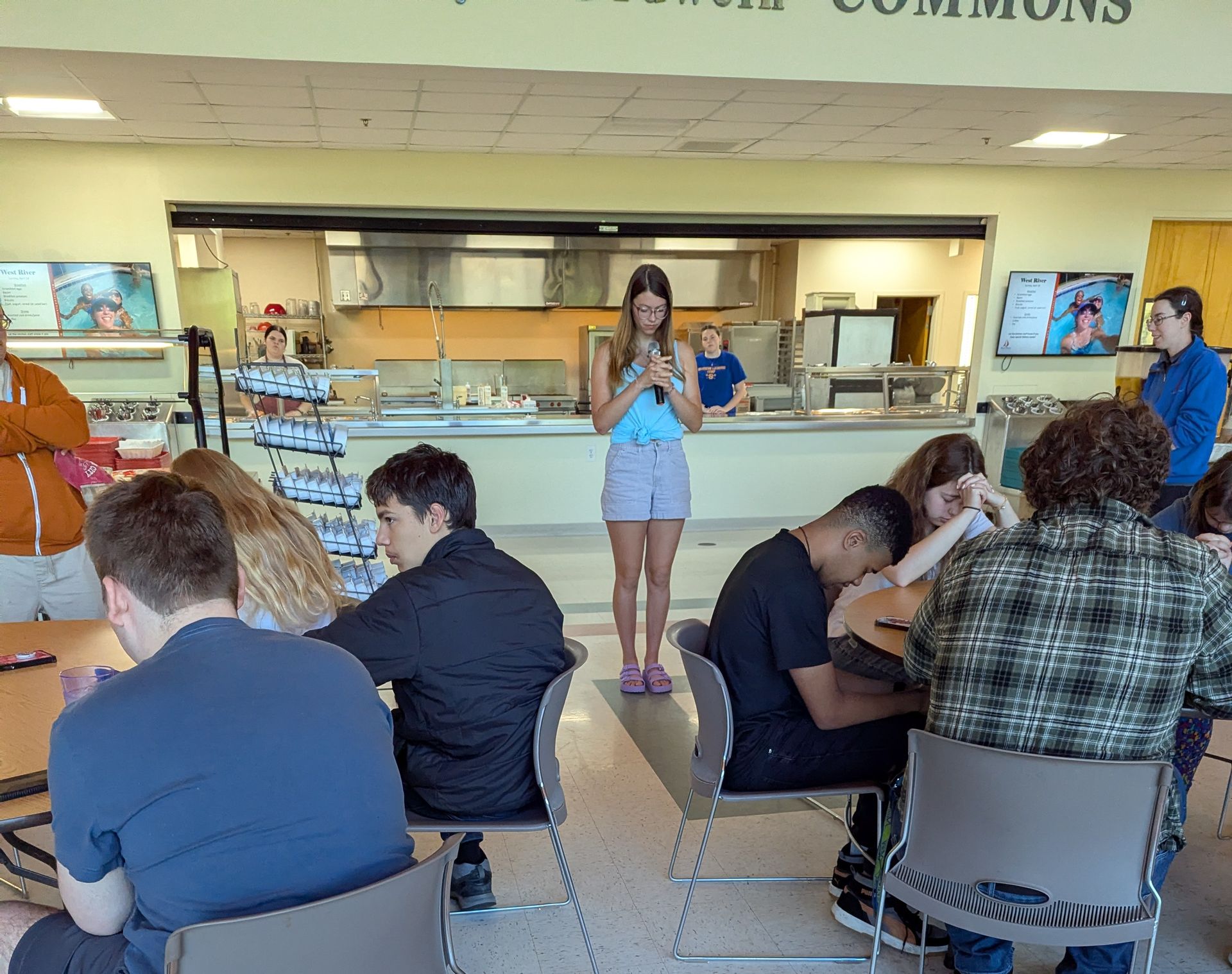 One youth saying grace  in a cafeteria while several youth bow their heads.