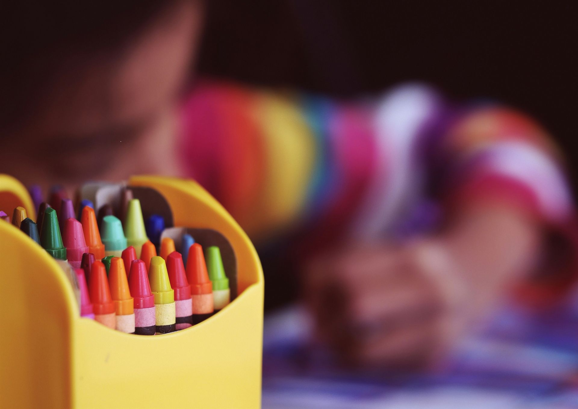Crayon holder with various colors in the foreground; child with a colorful shirt drawing in the background.