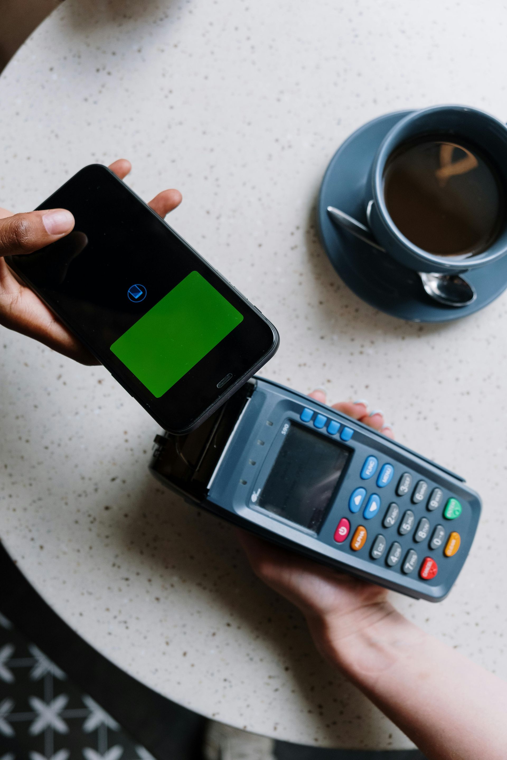 Person paying with phone at a point of sale terminal; green screen on the phone, coffee cup on the table.