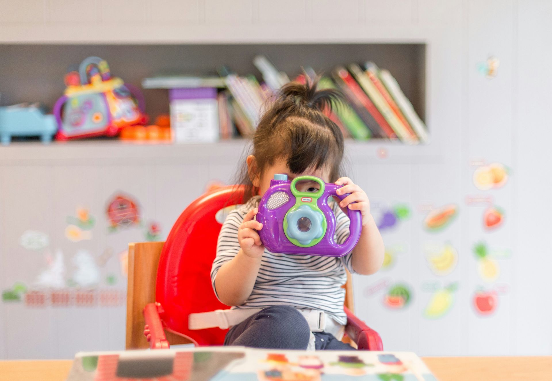 Young child in a high chair holding a purple toy camera, taking a picture.