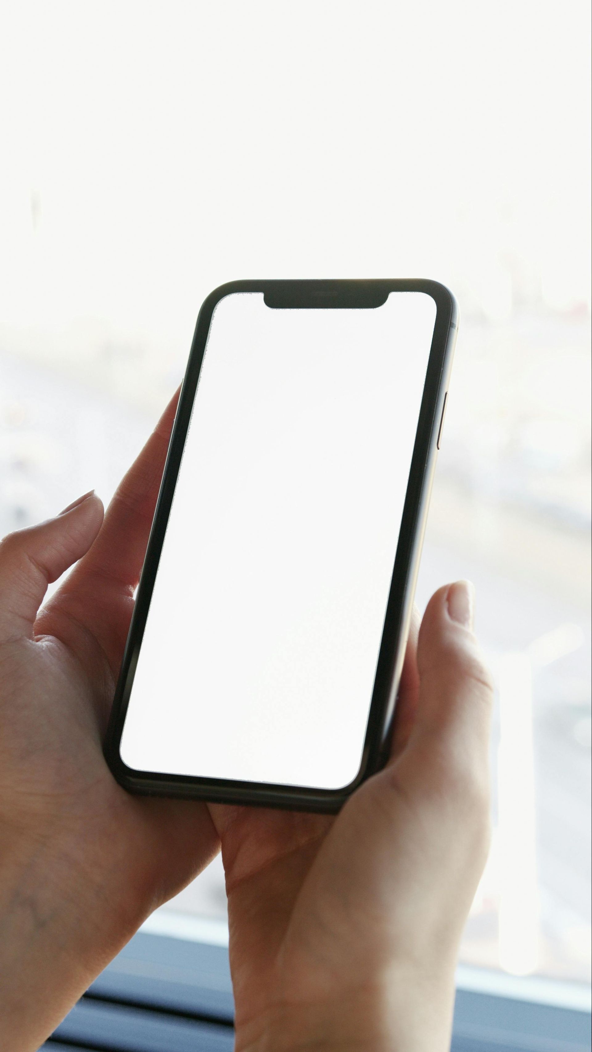 Hands holding a smartphone with a blank white screen, near a window.