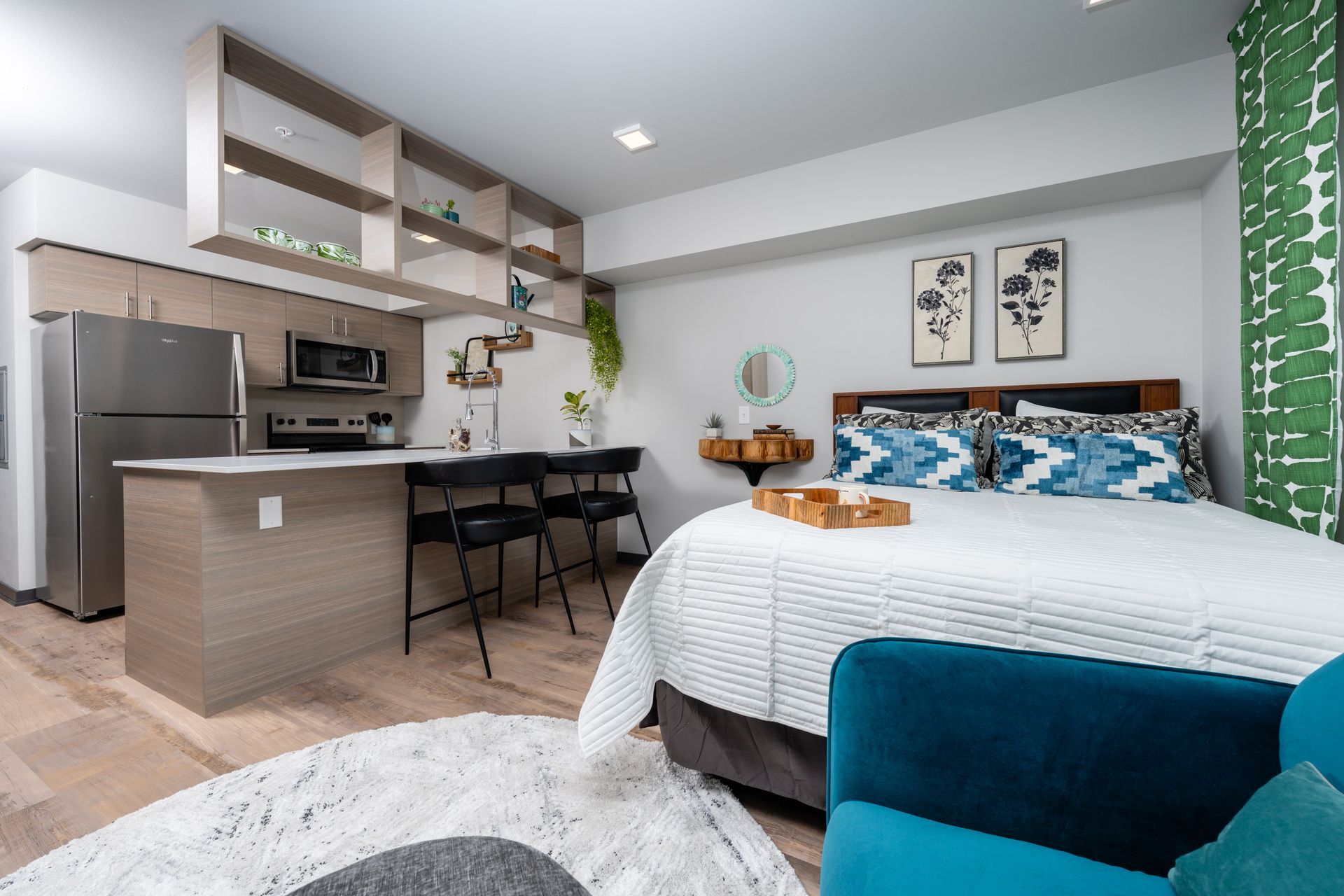 A modern studio apartment featuring a small kitchen, dining space with bar stools, and bed with blue accents, and a cozy rug.