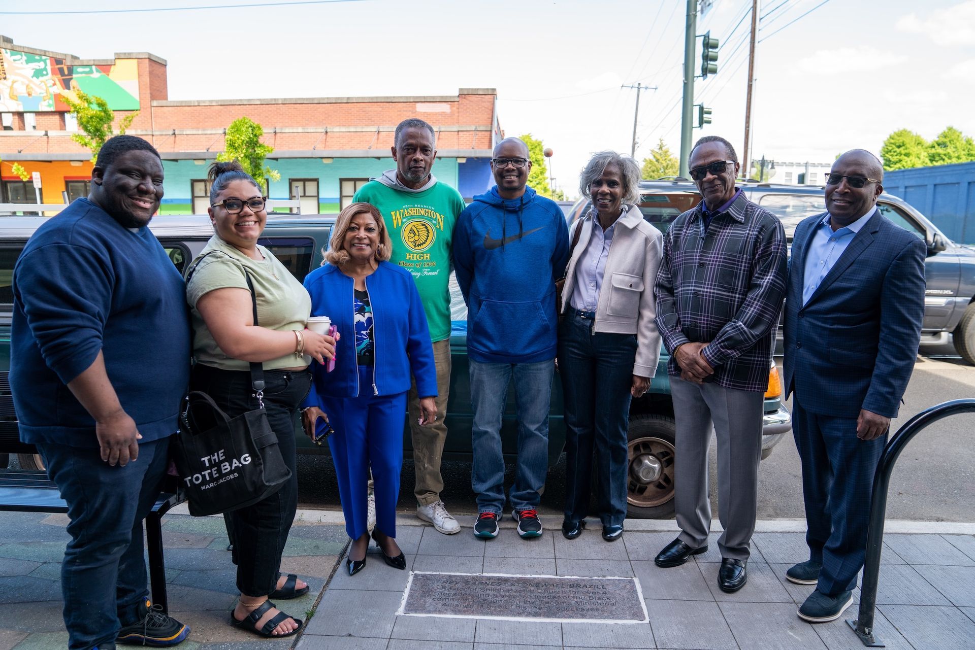 A group of nine people pose on a sidewalk in front of a building. Some are smiling; the setting appears to be outdoors.
