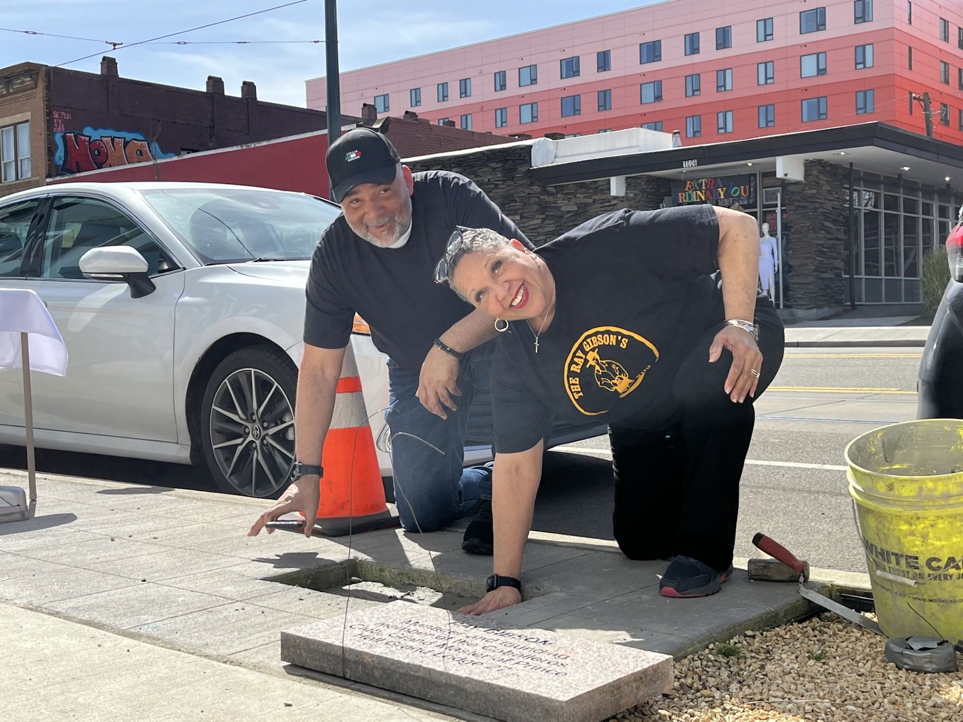 A couple presses hands on a concrete slab. Woman smiles, bending over, man kneels. Outside, cars and a building are visible.