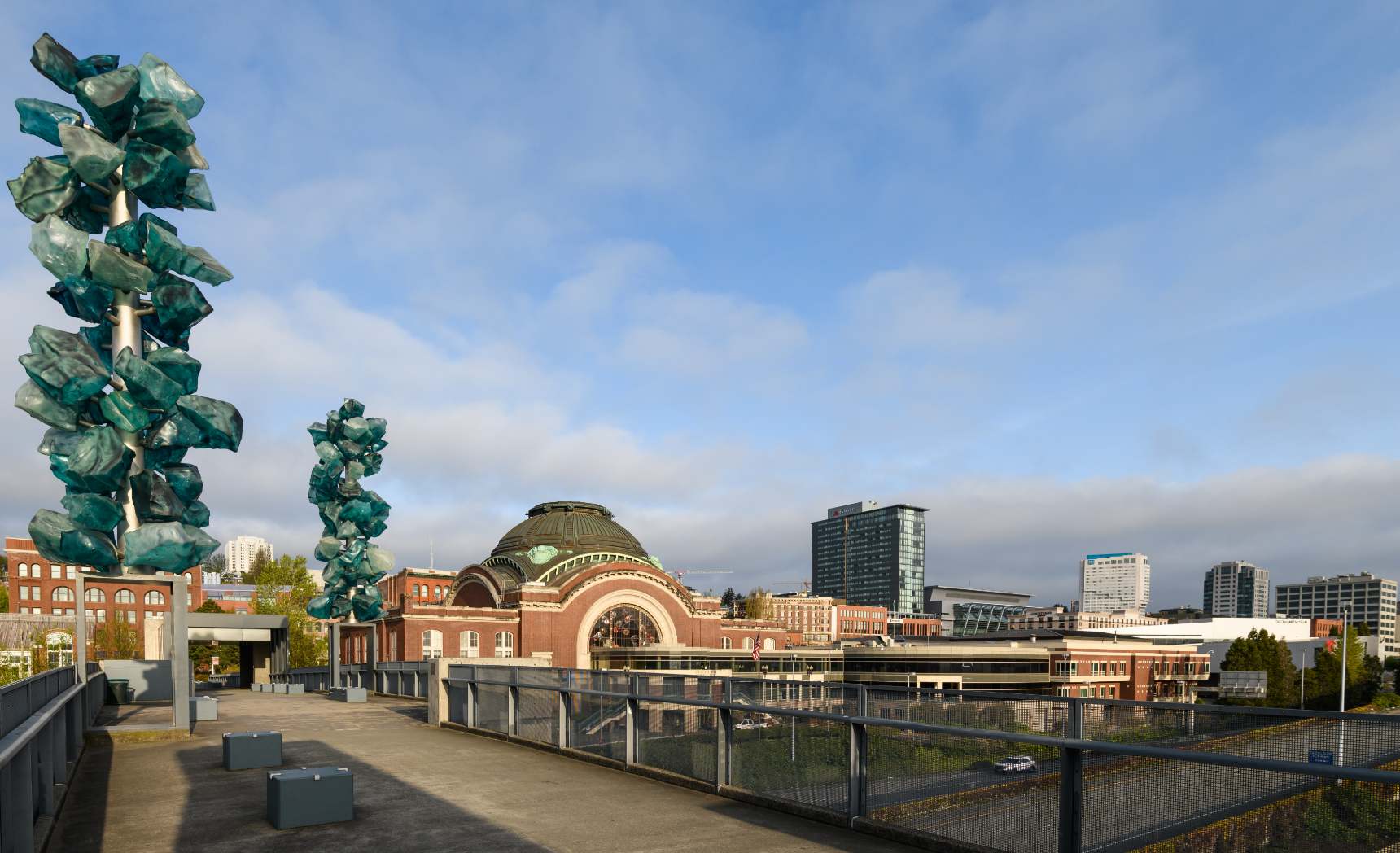 A cityscape includes art installations, a brick building, and modern high-rises under a cloudy sky.