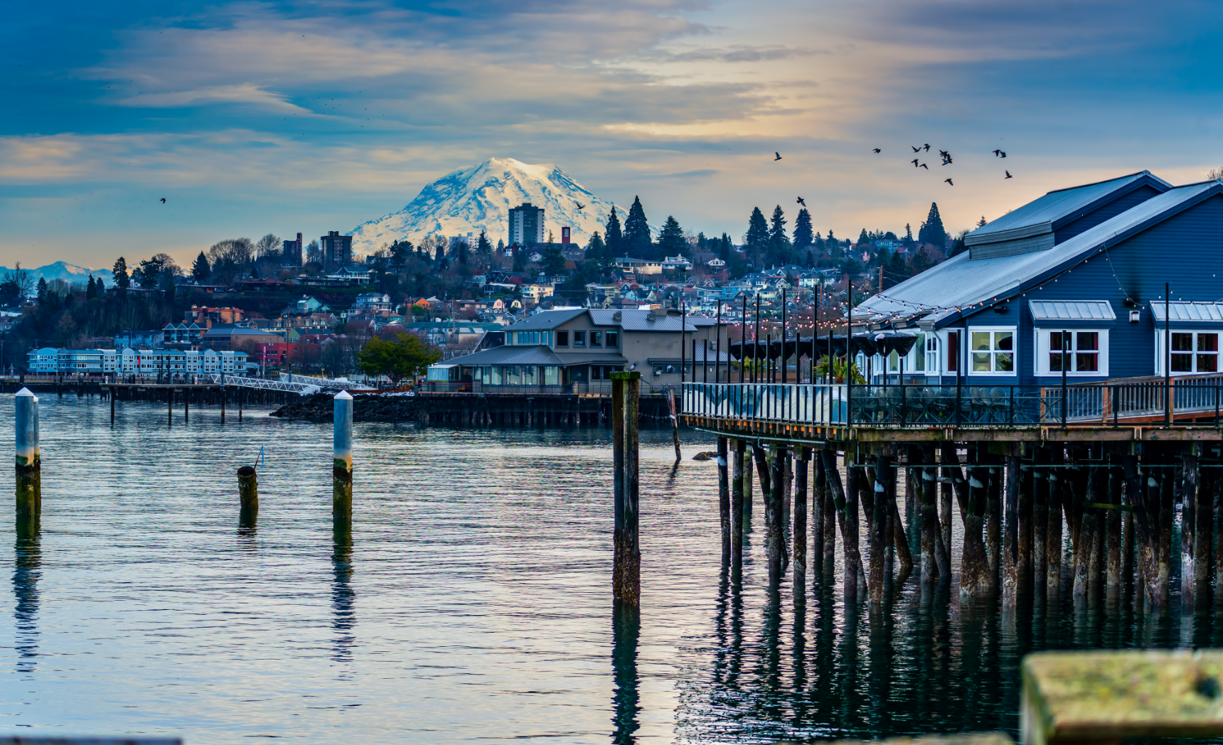 Waterfront view with buildings and pier, mountain backdrop, blue sky.