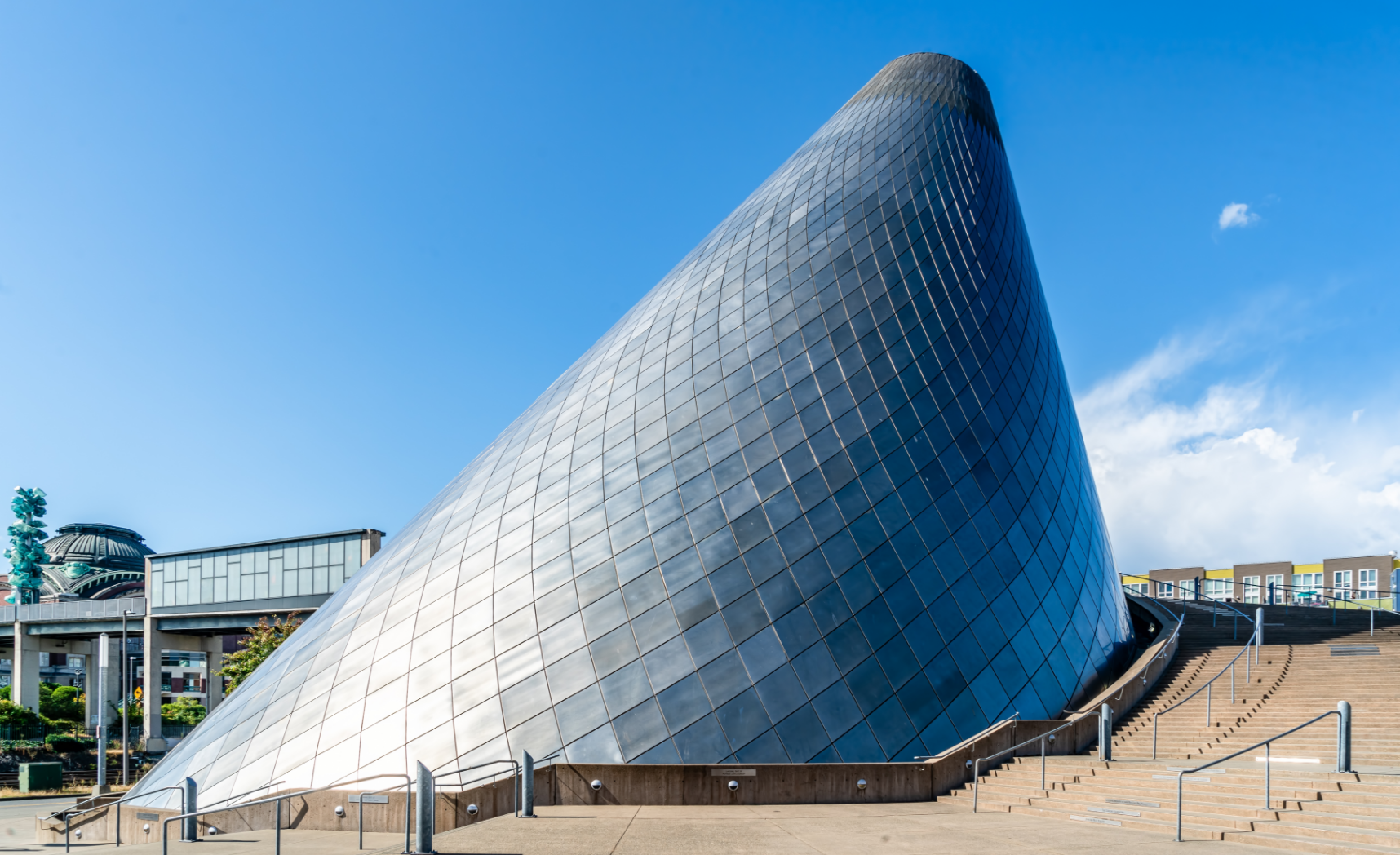 Conical, metallic building against a clear blue sky with a few clouds. Stone steps lead to the structure.