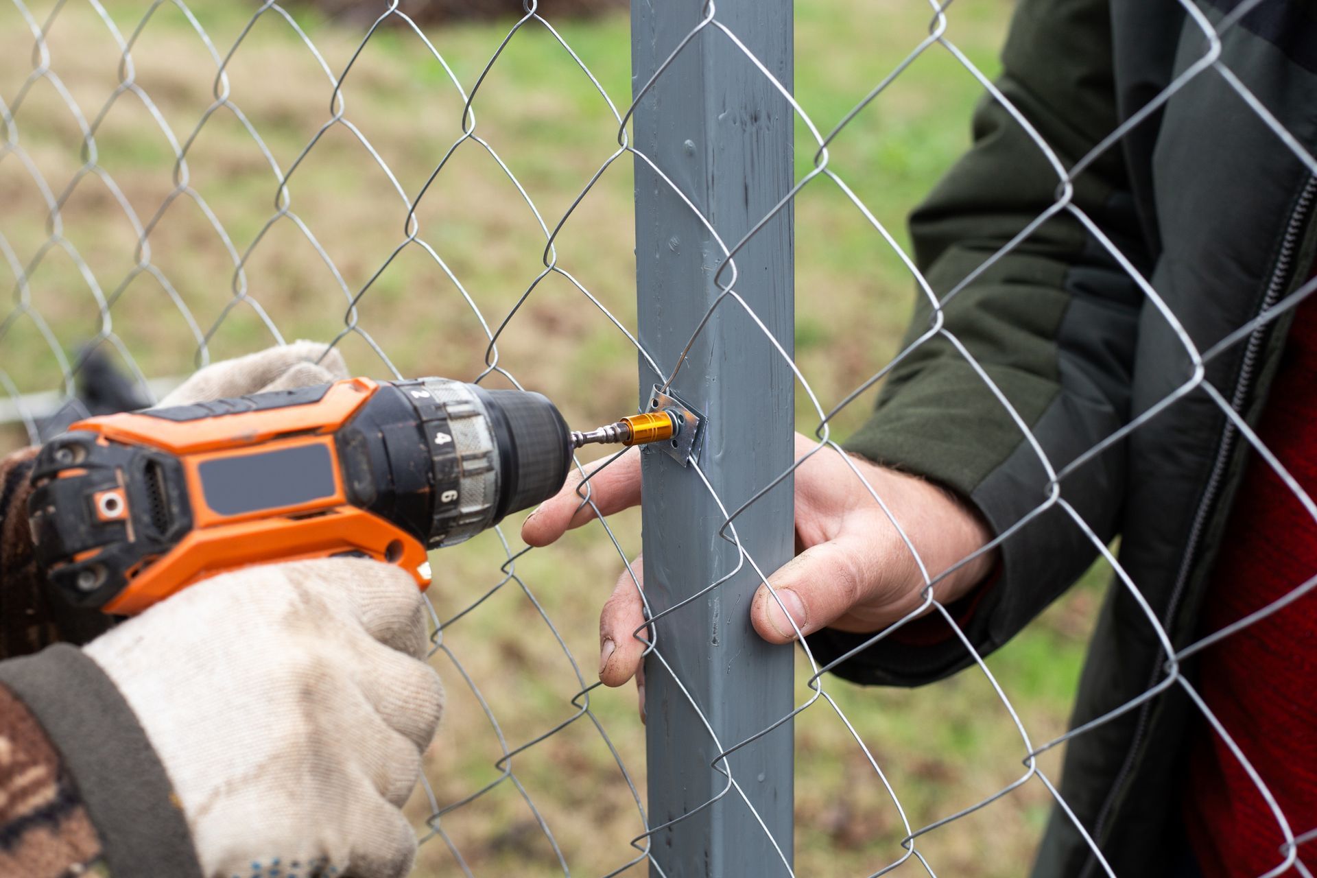 Person using a power drill to attach a metal post to a wire fence outdoors.
