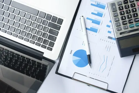 Laptop, business charts on clipboard, calculator, and pen on a white desk