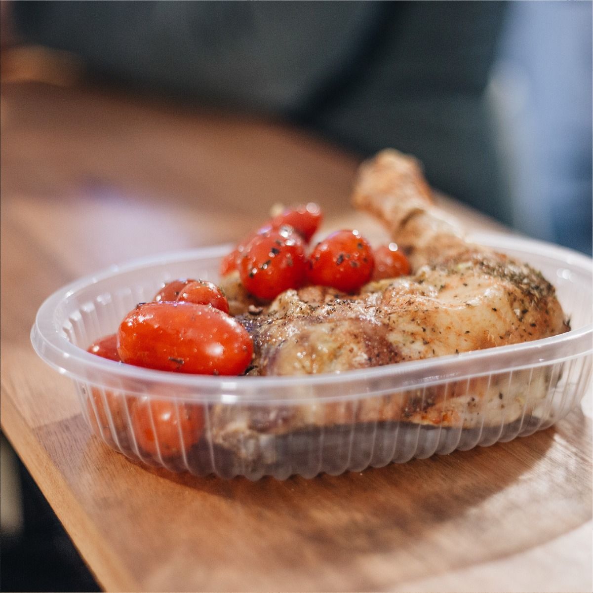Muslo de pollo y tomates asados ​​en un recipiente de plástico transparente sobre una mesa de madera.