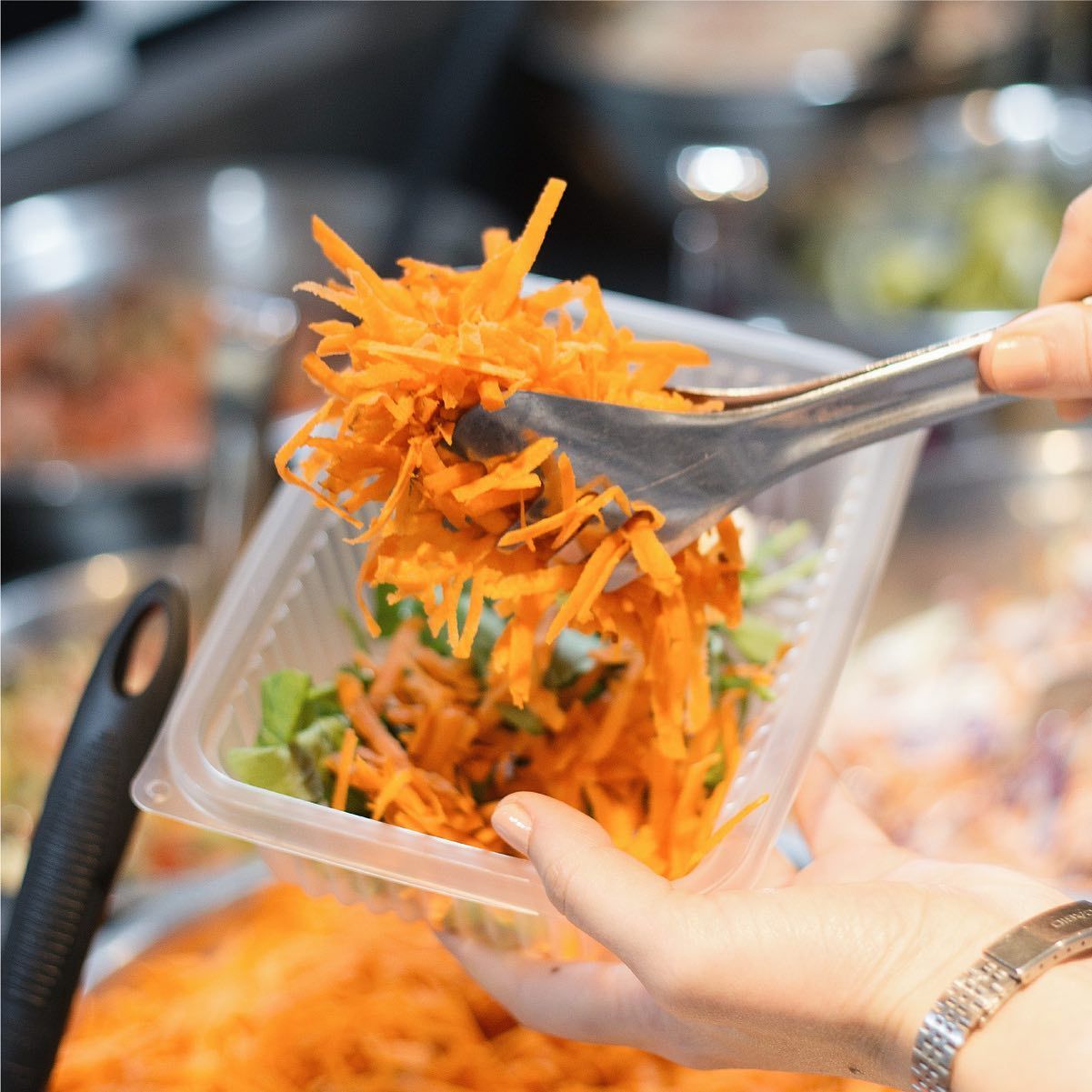 Persona que utiliza pinzas para añadir zanahorias ralladas a un recipiente de plástico con ensalada.