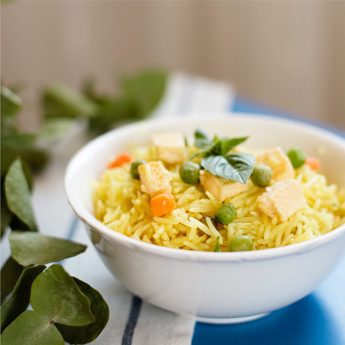 Arroz amarillo con tofu, guisantes y zanahorias en un bol blanco, adornado con albahaca.