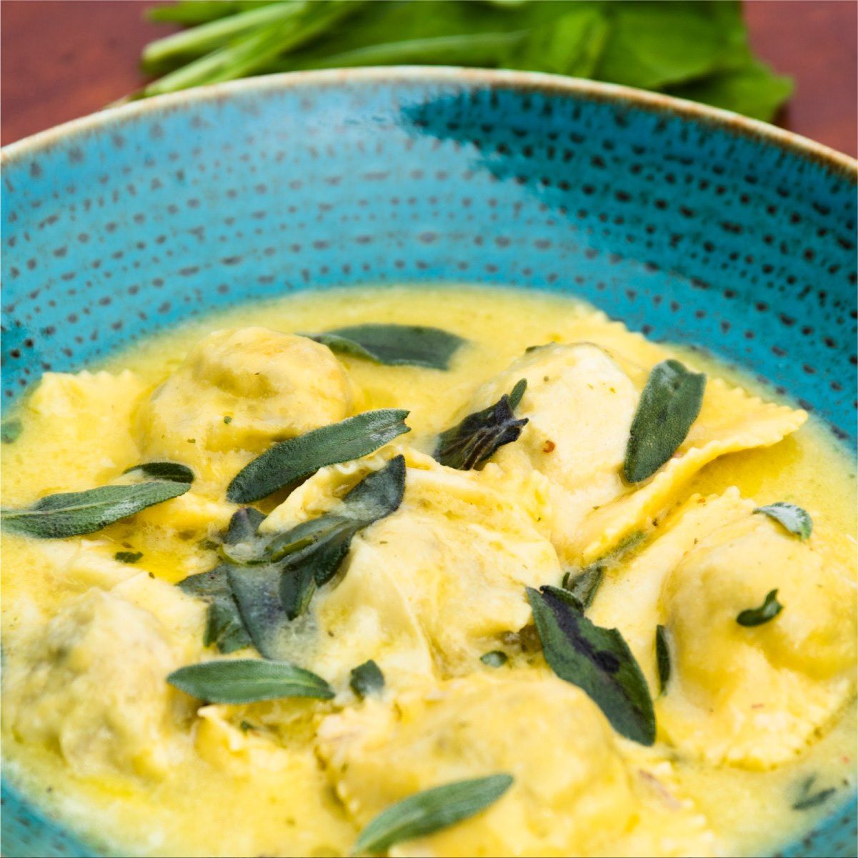 Raviolis en salsa amarilla cremosa, adornados con salvia, en un bol azul.