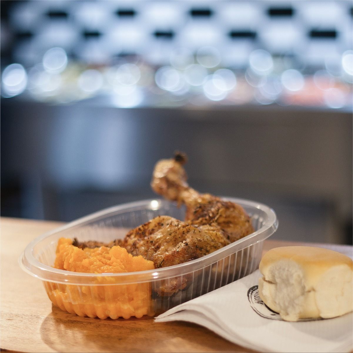 Pollo asado y puré de batatas en un recipiente de plástico, con un panecillo sobre una servilleta.