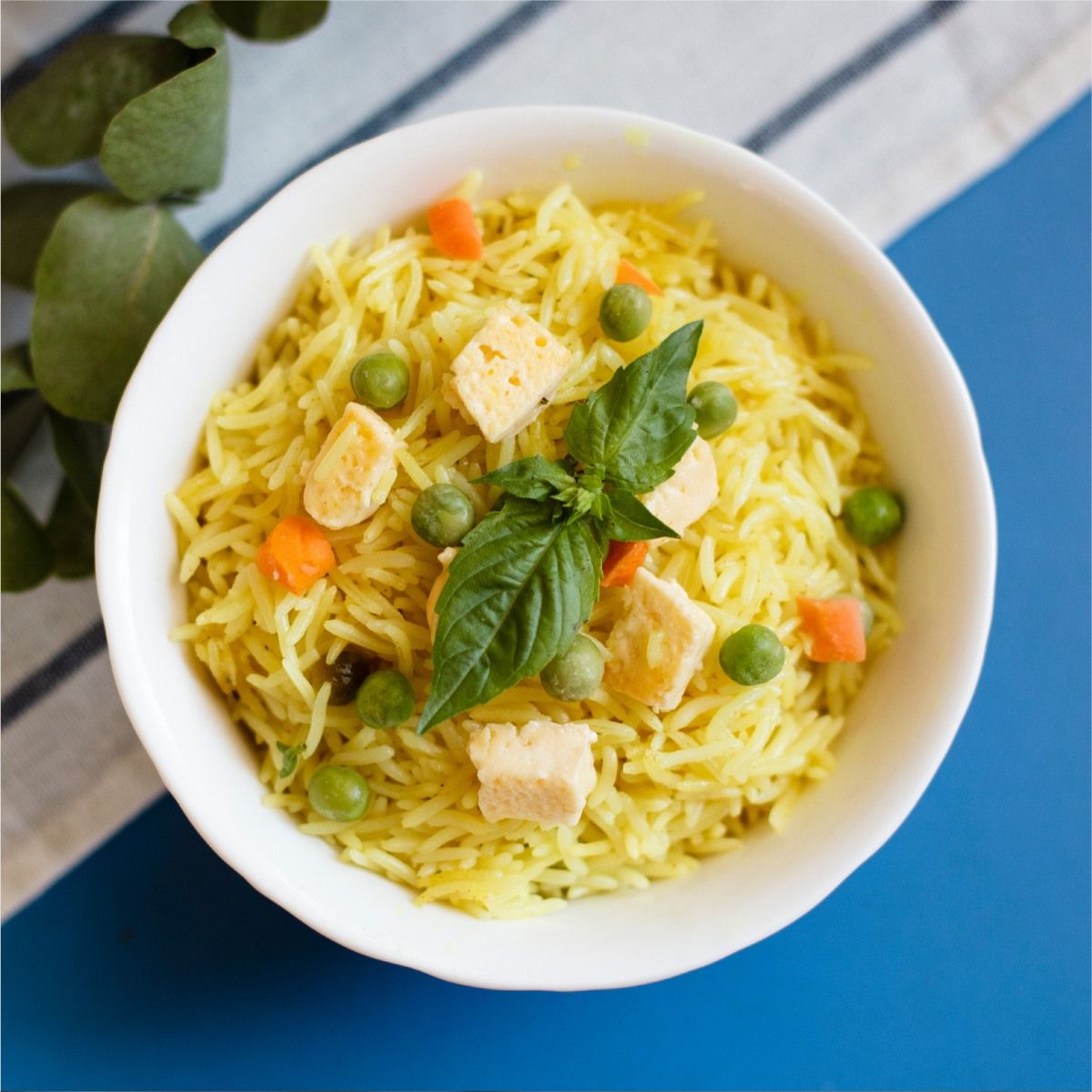 Tazón de arroz amarillo con tofu, guisantes, zanahorias y albahaca sobre un fondo azul.