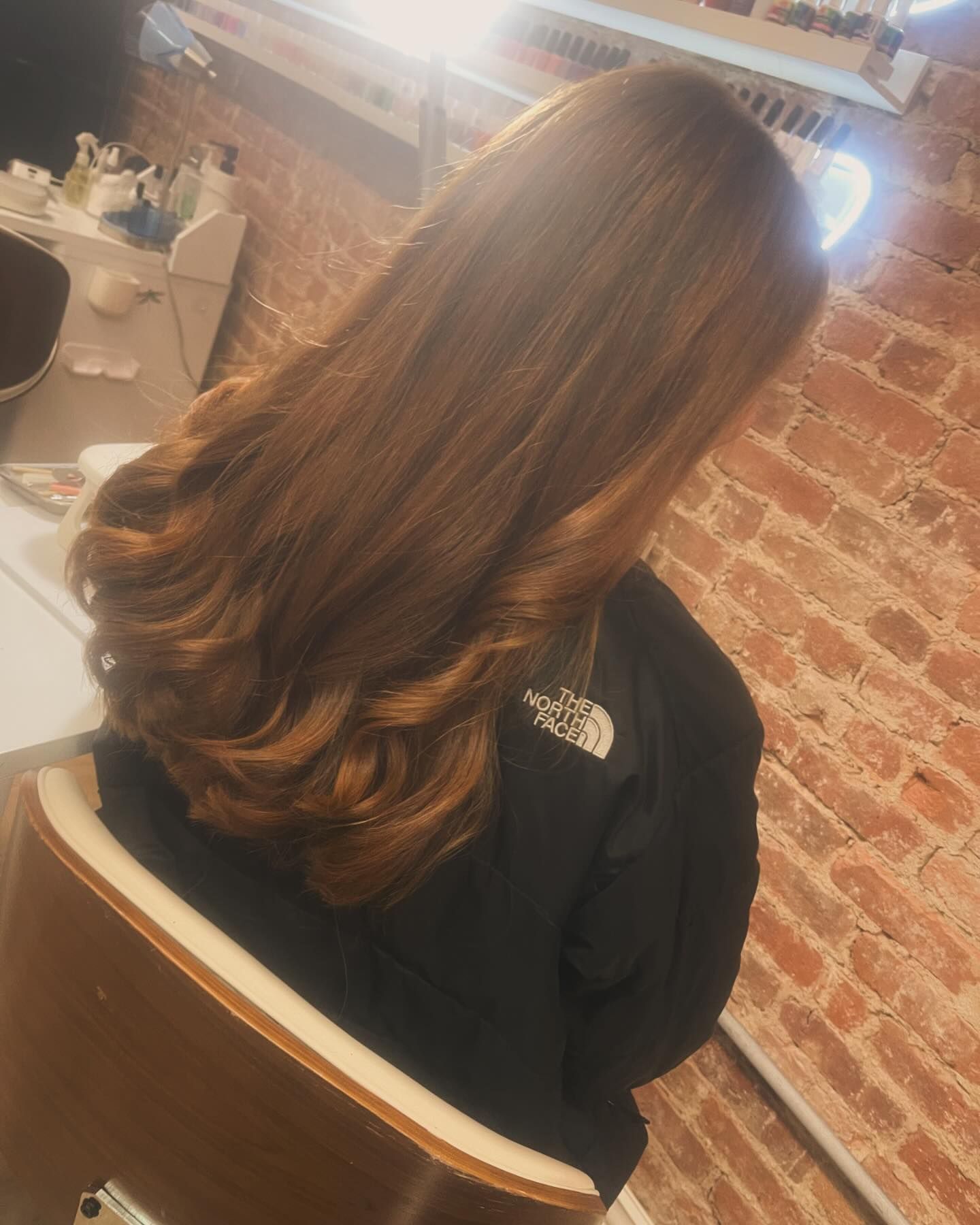 A woman with long brown hair is sitting in a chair in front of a brick wall.